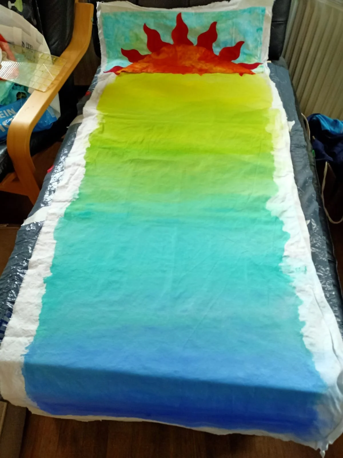 Hand-painted fabric with a design of a sunset over a body of water, featuring a large red and orange sun with flames, and a gradient sky transitioning from blue to green and yellow.