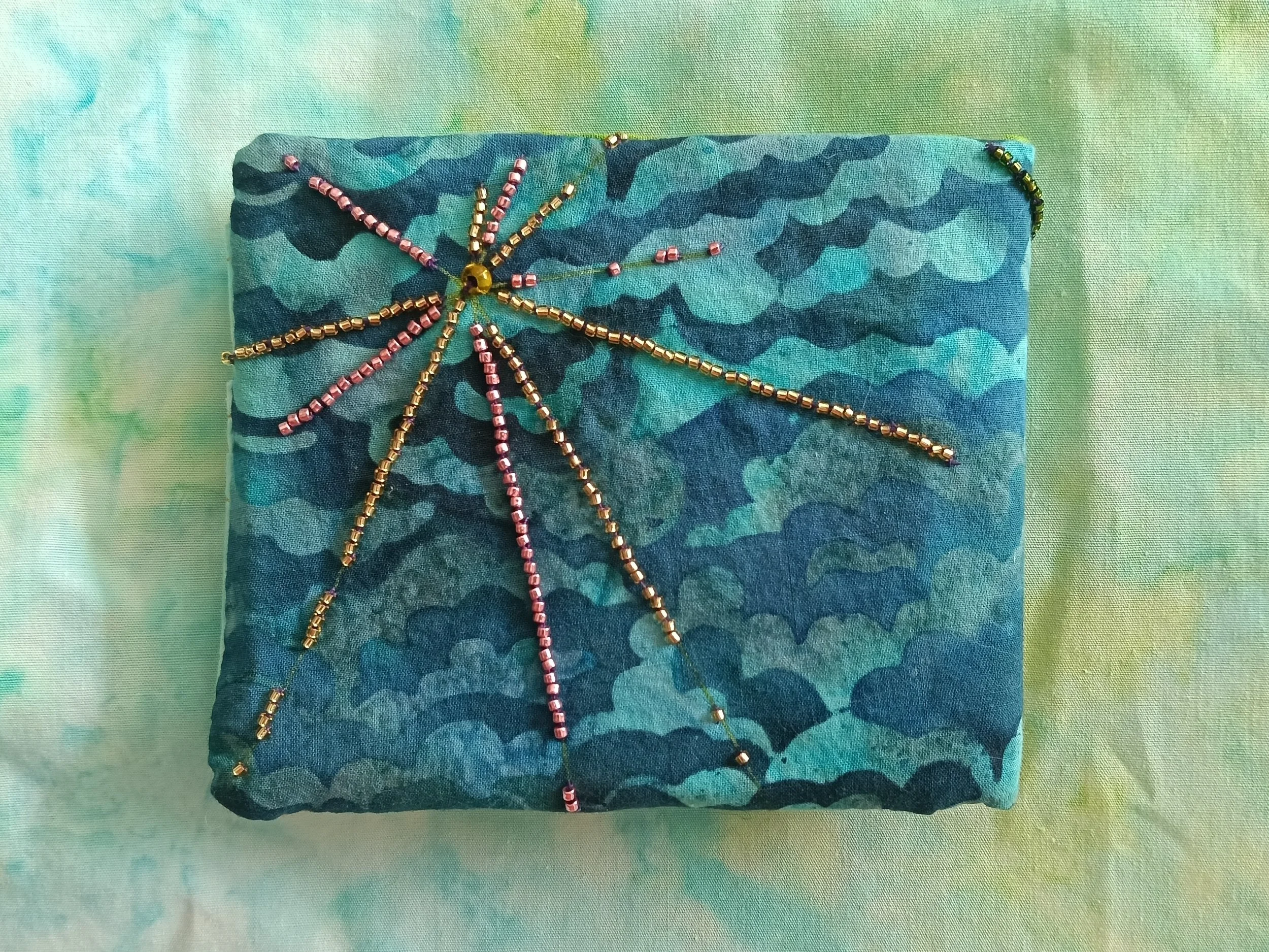Colorful beaded jewelry with pink and gold beads arranged in a star-like pattern on a teal fabric background with a greenish pastel surface.