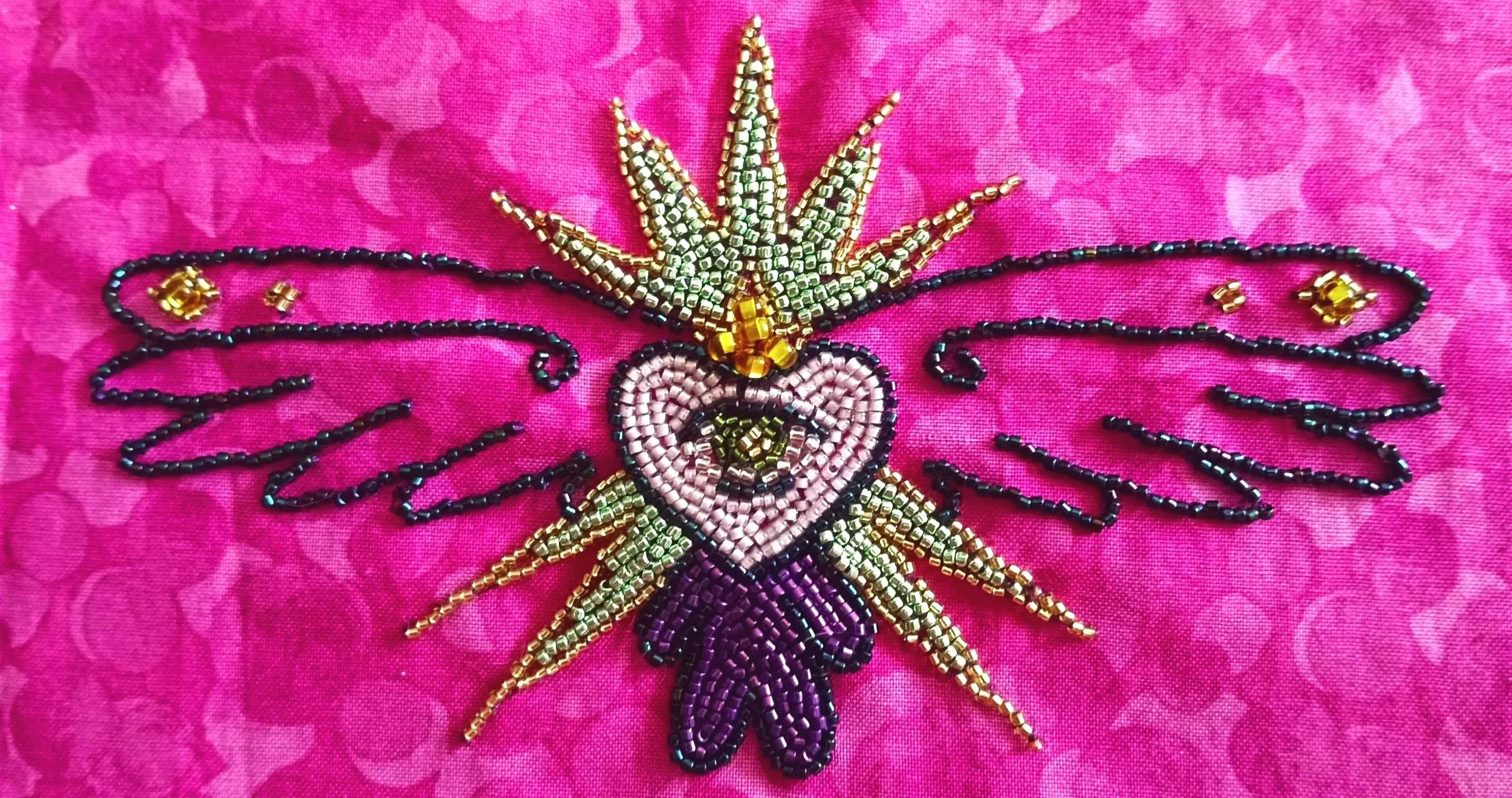 Beaded embroidery of a fairy with wings, holding a heart, on pink fabric.
