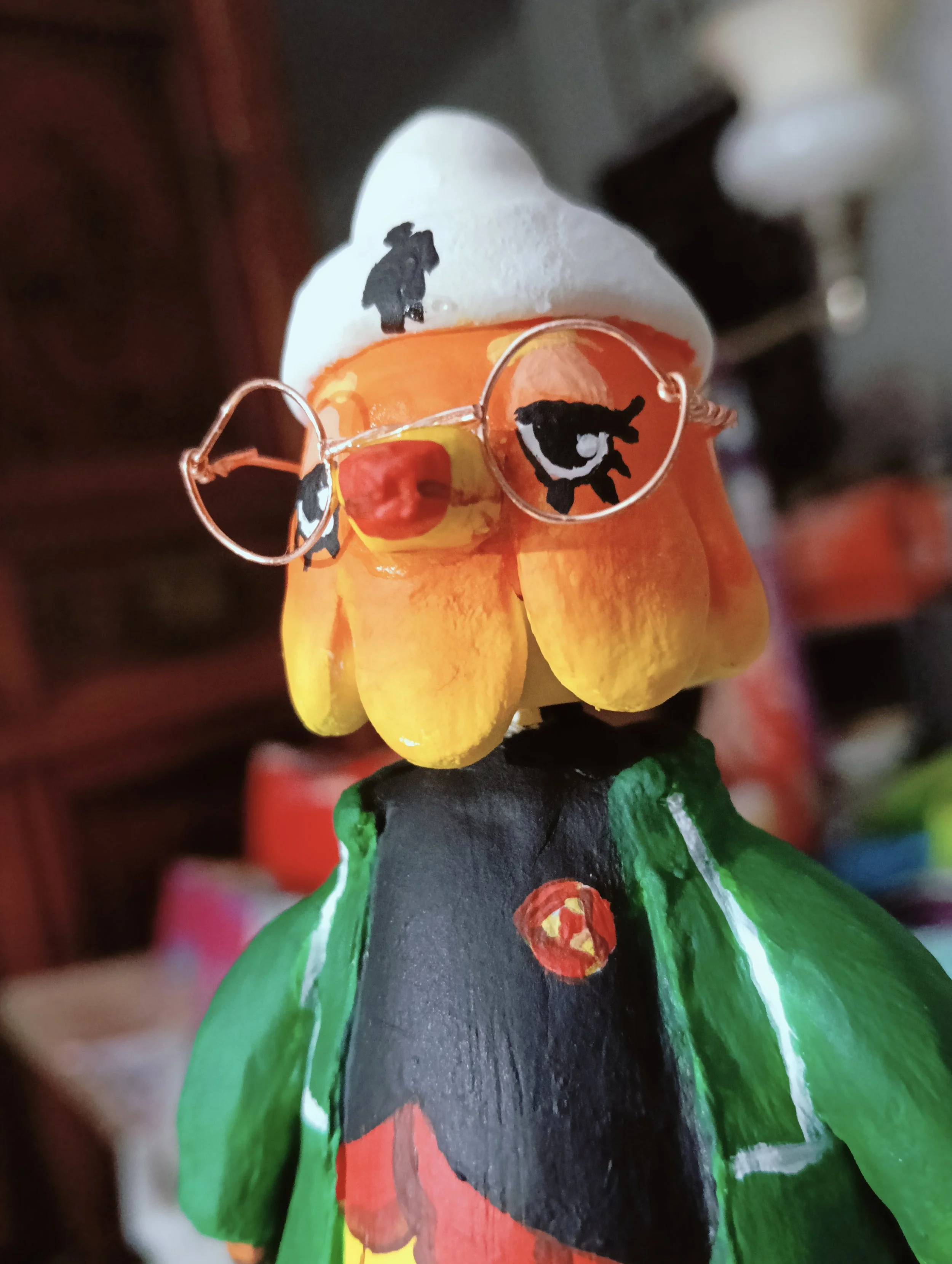 Close-up of a painted bird figurine wearing glasses, with black hair, a red nose, and a costume with green, red, and yellow colors.