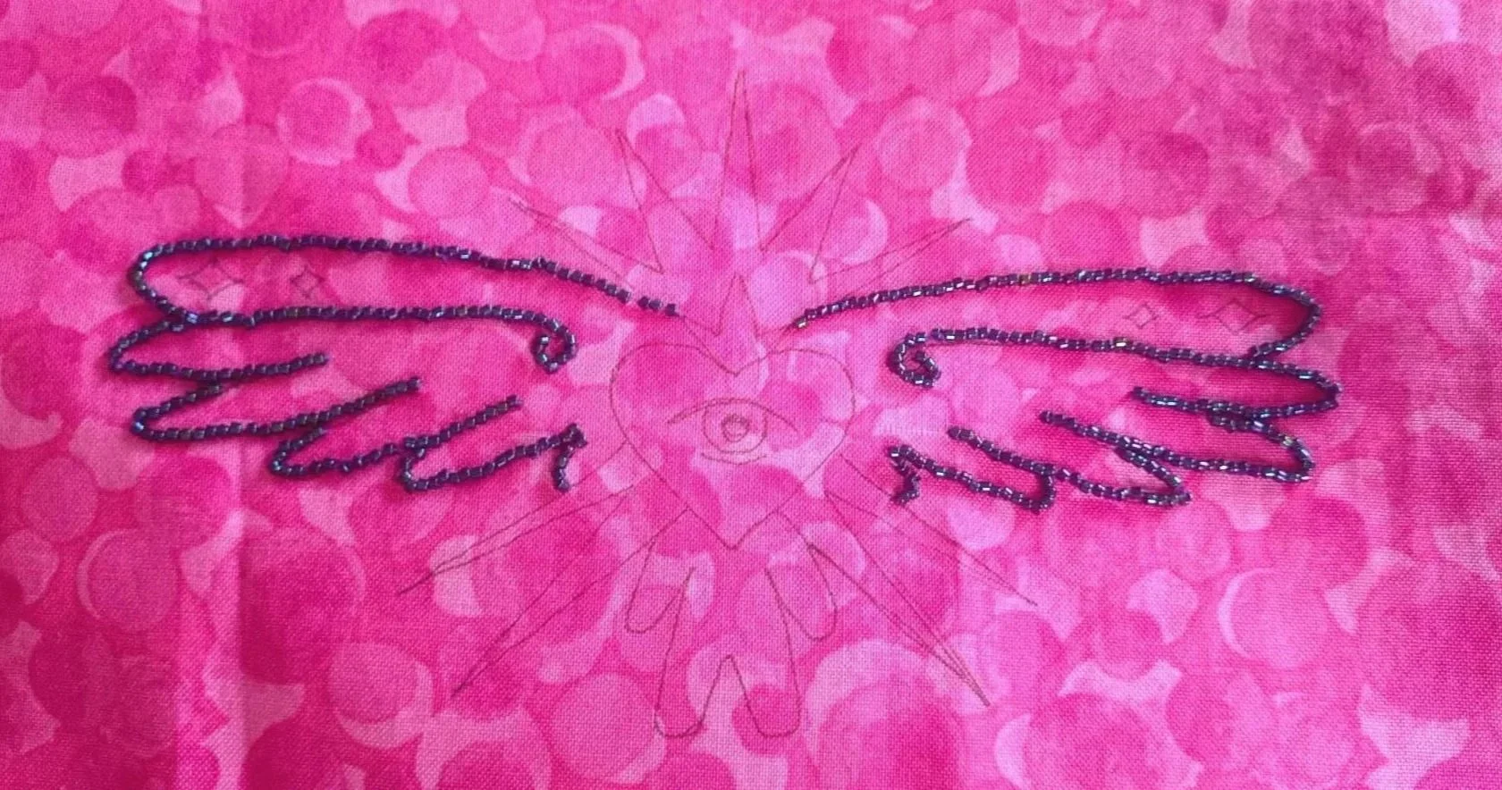 Pink fabric with a printed heart and stars design, featuring a wired outline of angel wings embroidered with black beads and sequins.
