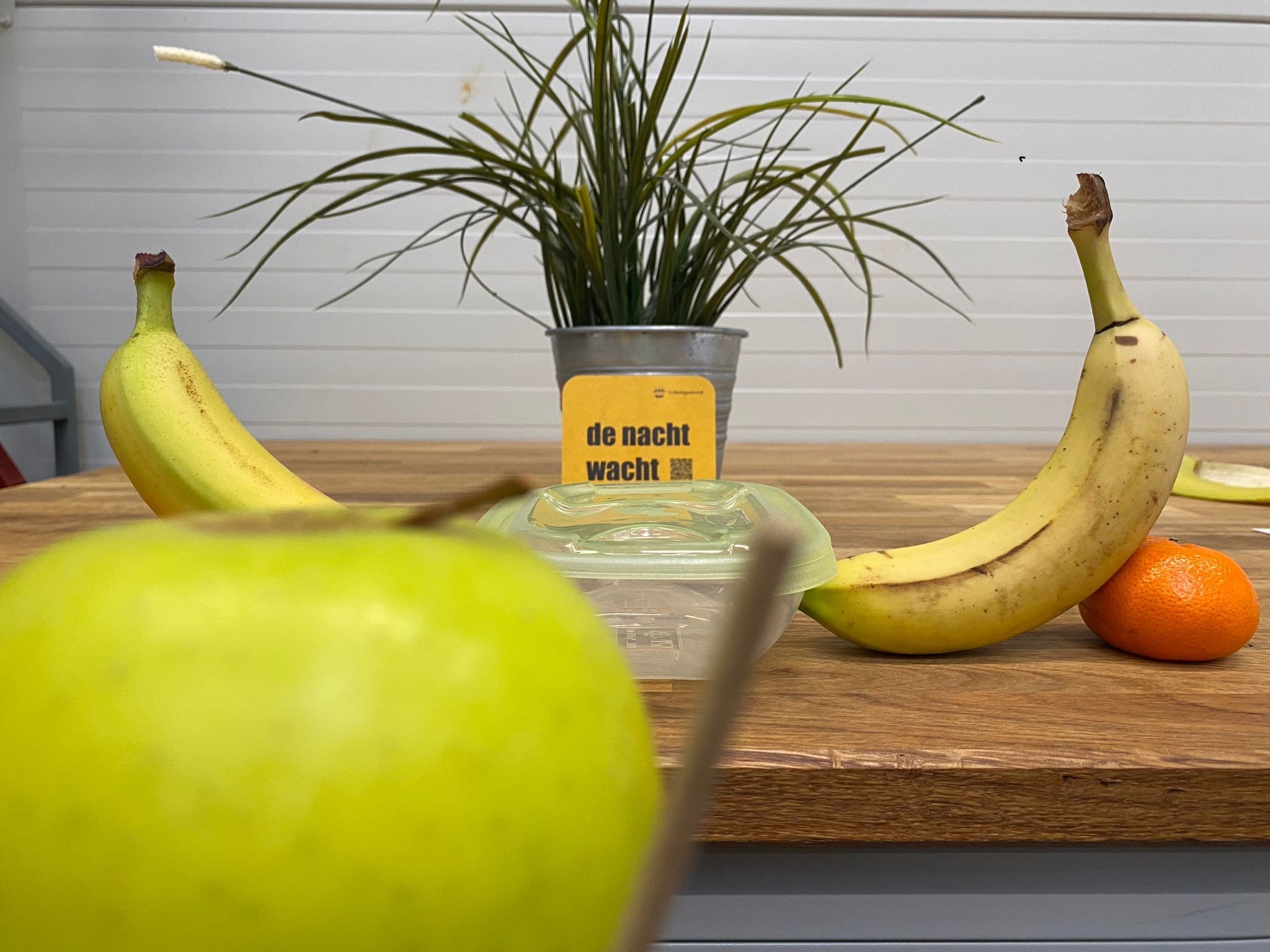 A photograph of a decorative arrangement on a wooden table. The arrangement includes two bananas, one on each side, a yellow box with black and yellow text in the center, a gray pot with a green plant behind the box, and two oranges at the bottom right corner. The background features a white wall with horizontal siding.