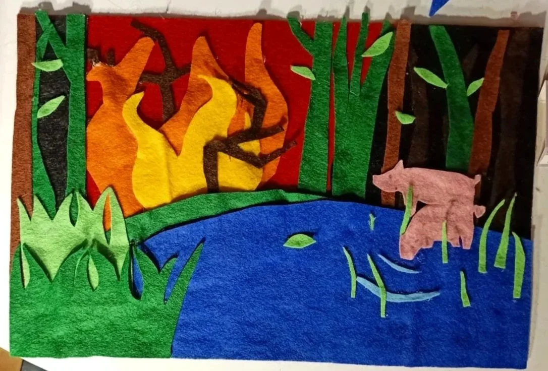 Felt art scene of a forest with a pond, pink pig, trees, and a fire in the background.