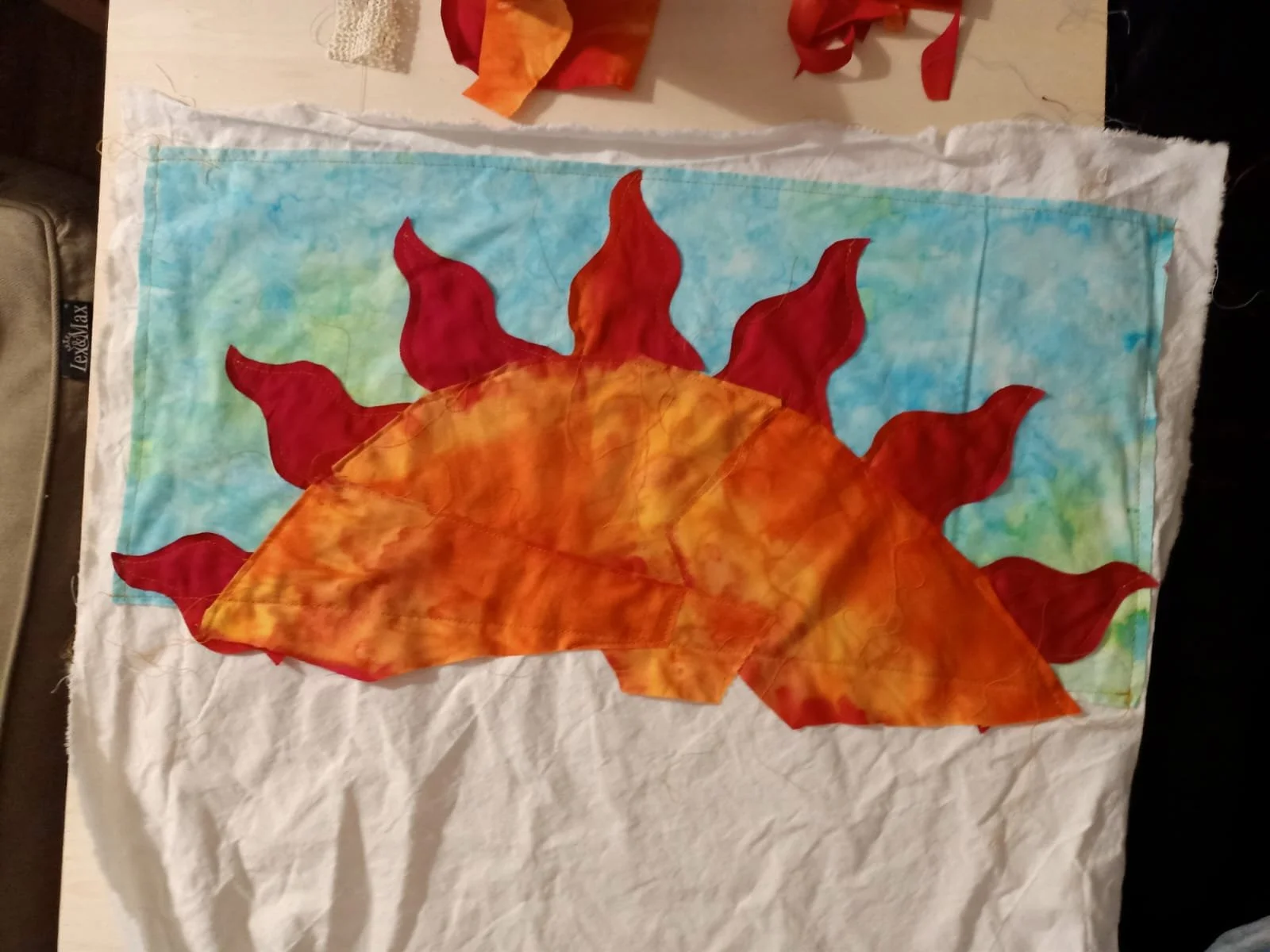 A quilt with a design of a large sun made of autumn-colored fabrics, set against a blue sky background with green accents.