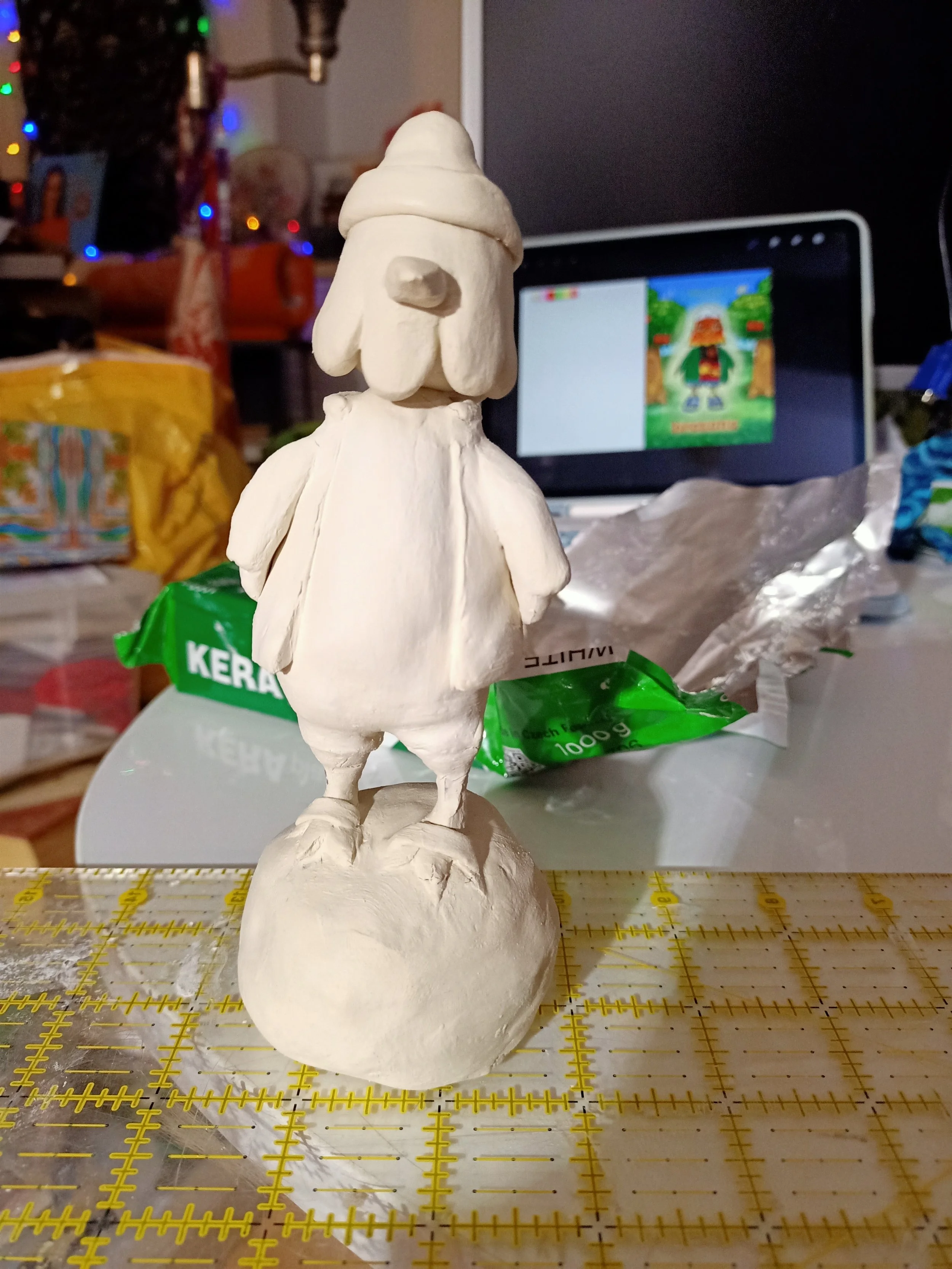 A white clay figure of a fictional character with a large nose, wearing a hat and a cloak, standing on a round base on a cutting mat. Background shows a computer monitor with colorful art, some clutter, and decorations.
