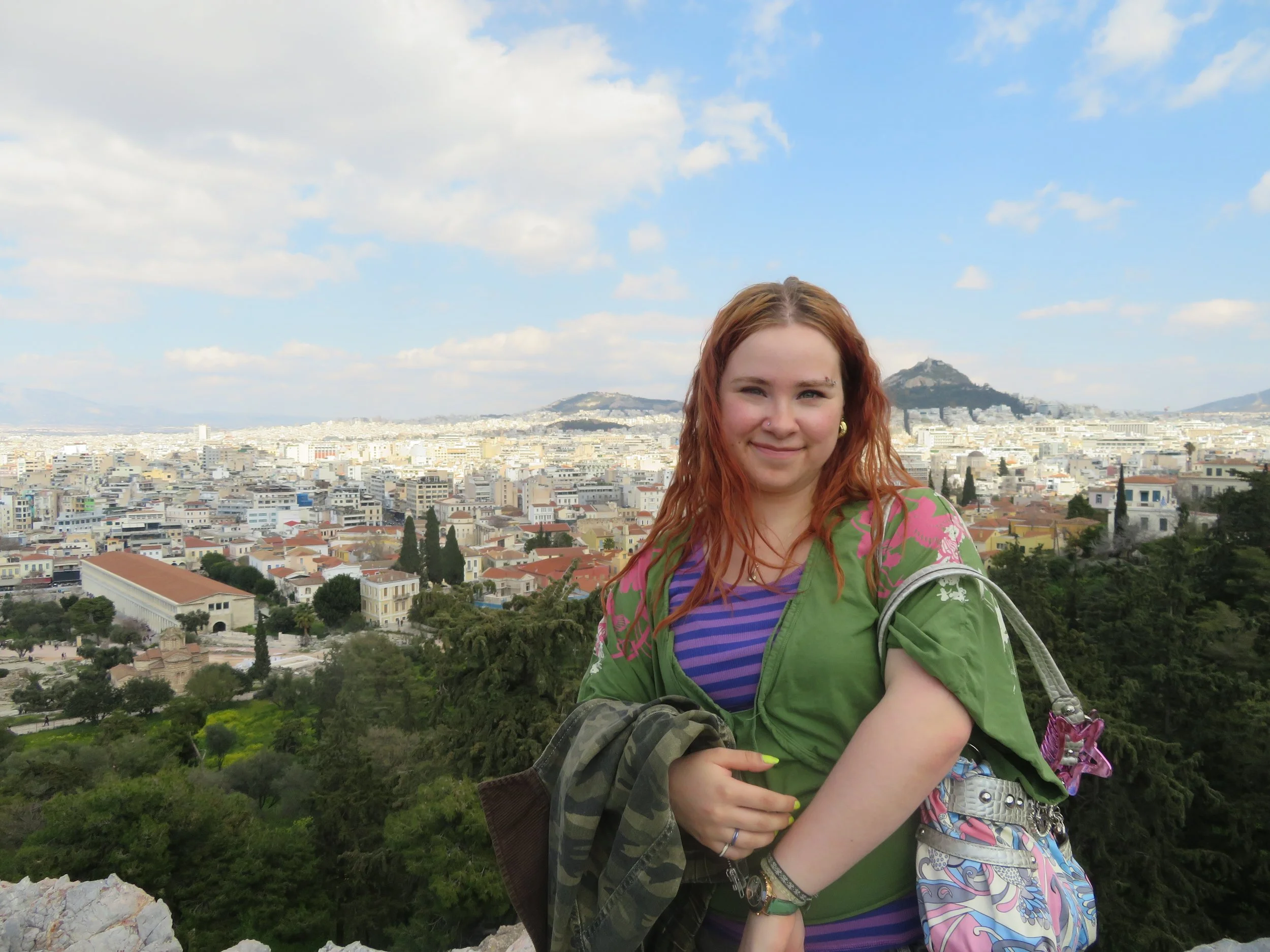 A young woman with red hair and a nose piercing wearing a green jacket and a multicolored striped shirt, standing outdoors with a cityscape and hills in the background under a partly cloudy sky.