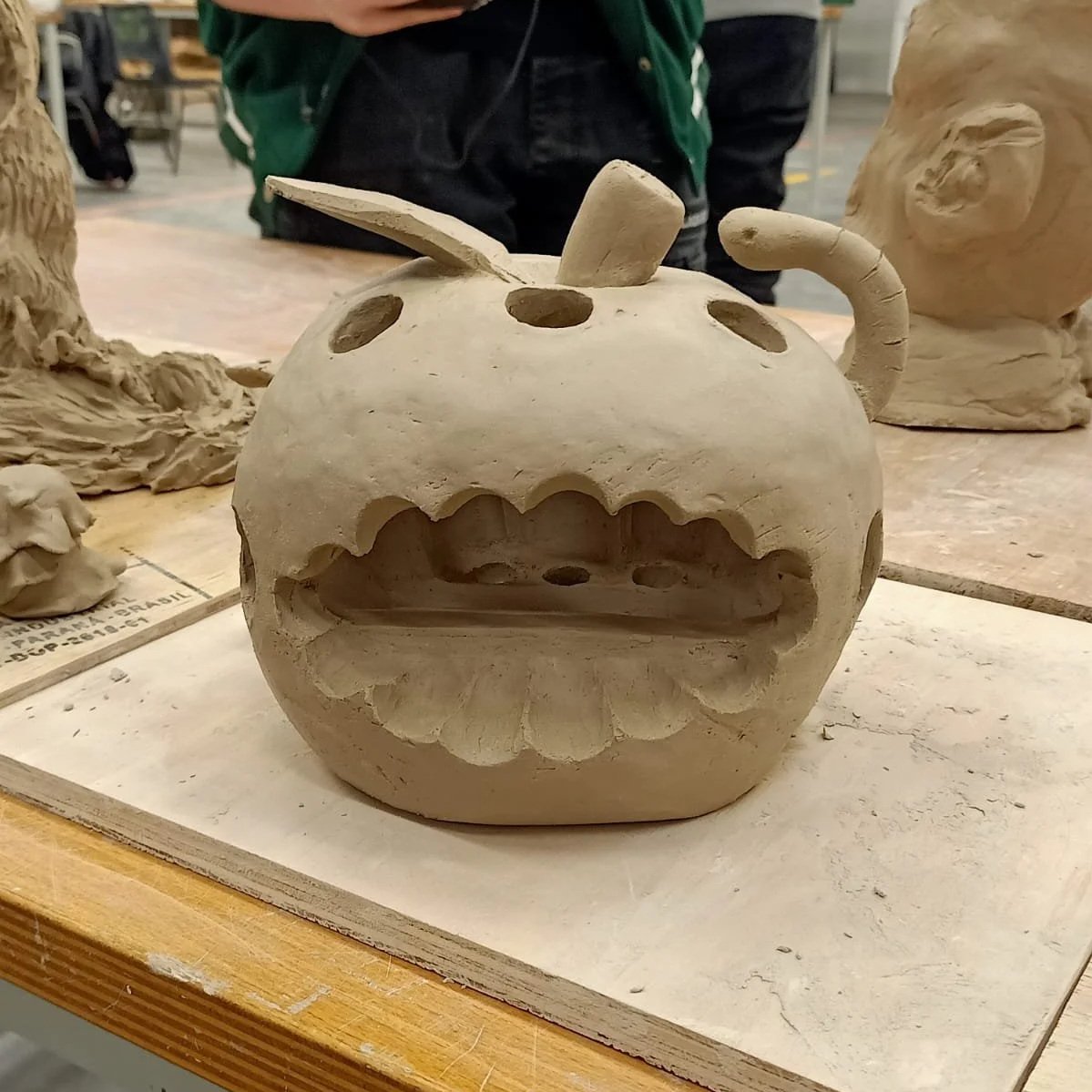 A small, unglazed clay sculpture of an apple with a carved face, featuring an open mouth with jagged teeth, circular eyes, a tiny nose, a small cylindrical stem, and a curved handle, placed on a wooden worktable in a ceramics studio.