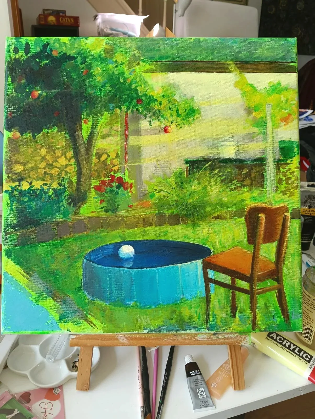 A bright, colorful painting of a lush garden scene with a tree bearing red fruit, a small blue pond with a white ball floating inside, a wooden chair, and various green plants and bushes, set against a house and blue sky in the background.