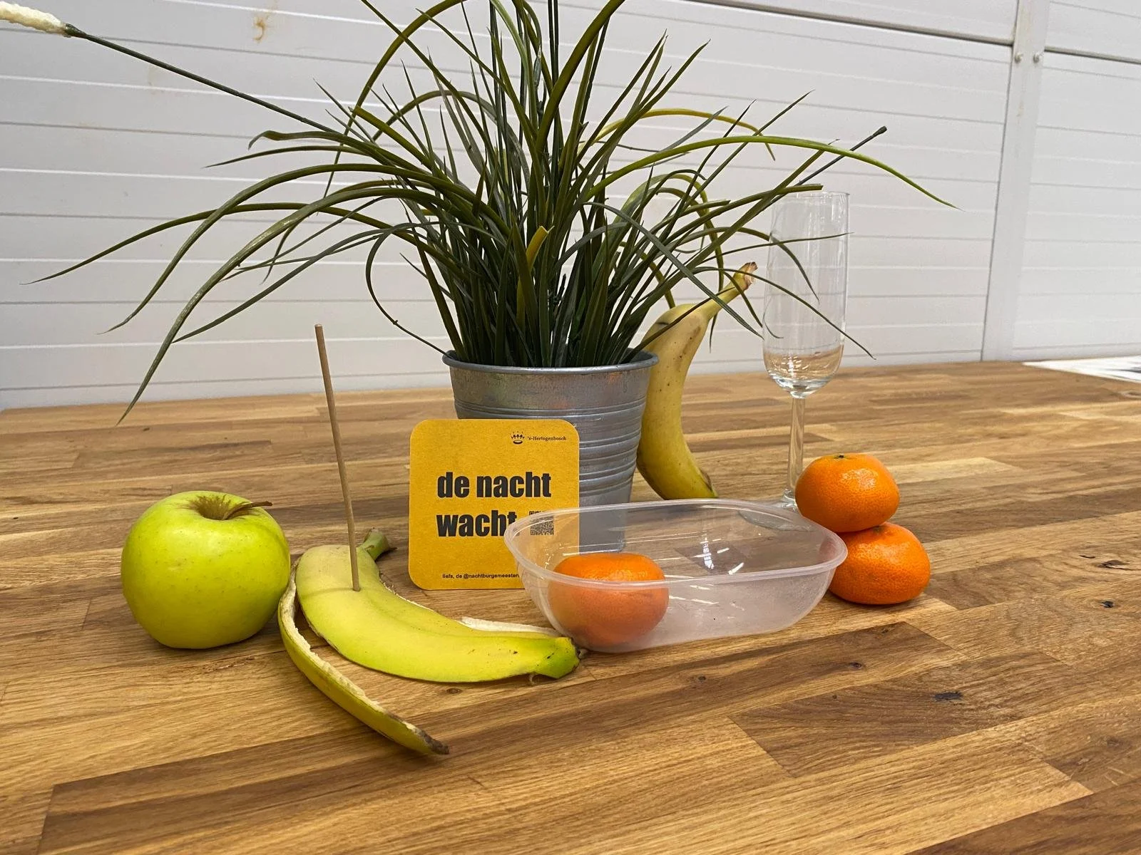 A wooden table with an apple, bananas, oranges, a glass of water, a potted plant, and a yellow sign with black text in Dutch.