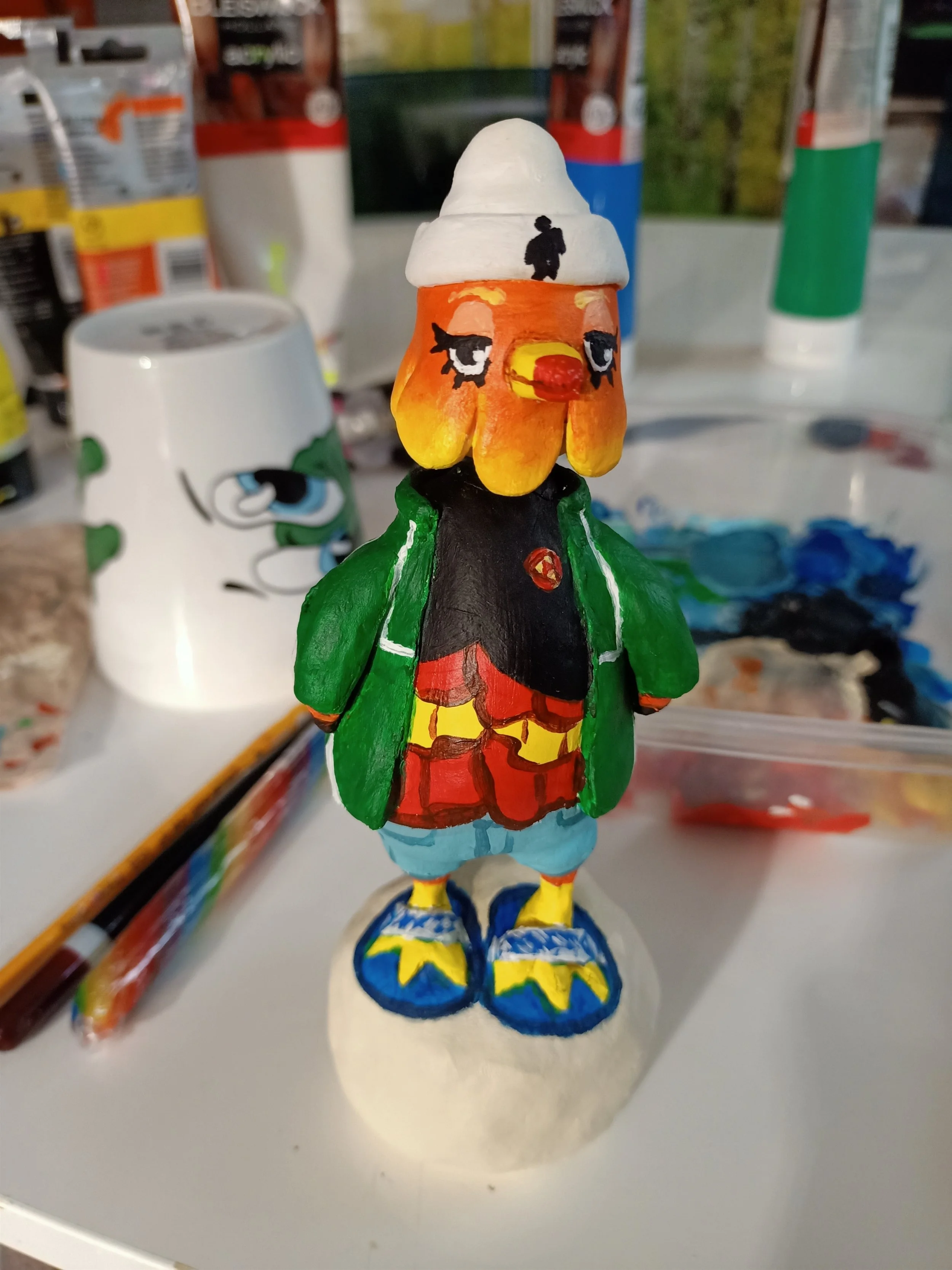 Hand-painted figurine of a chicken wearing a white hat with a small person figure, a black jacket, red shorts, yellow and blue shoes, standing on a colorful base.