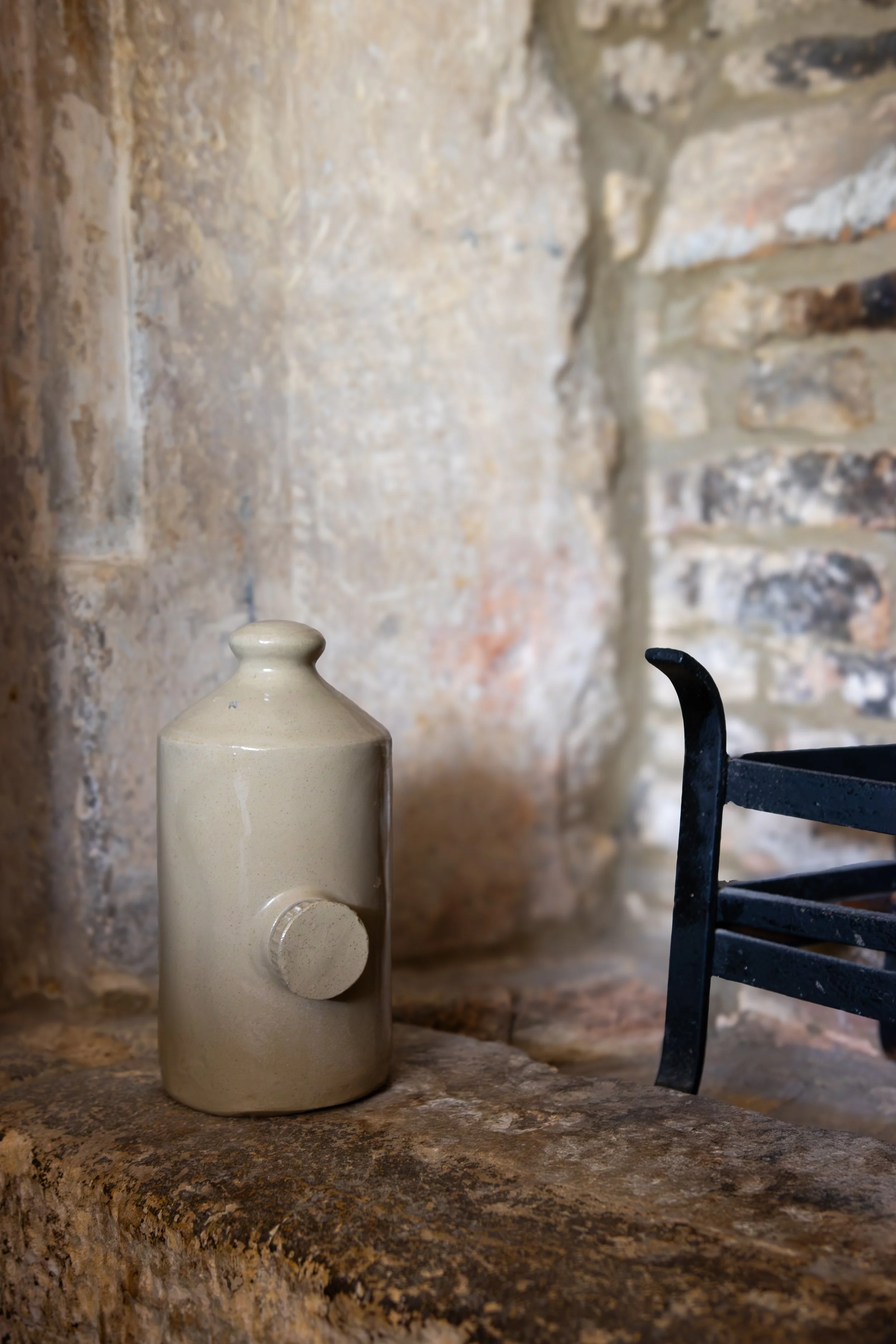 A beige ceramic bottle with a round stopper placed on a rough stone surface, in front of a brick wall background with a black metal curtain rod or pipe to the right. The Lamb Inn. 