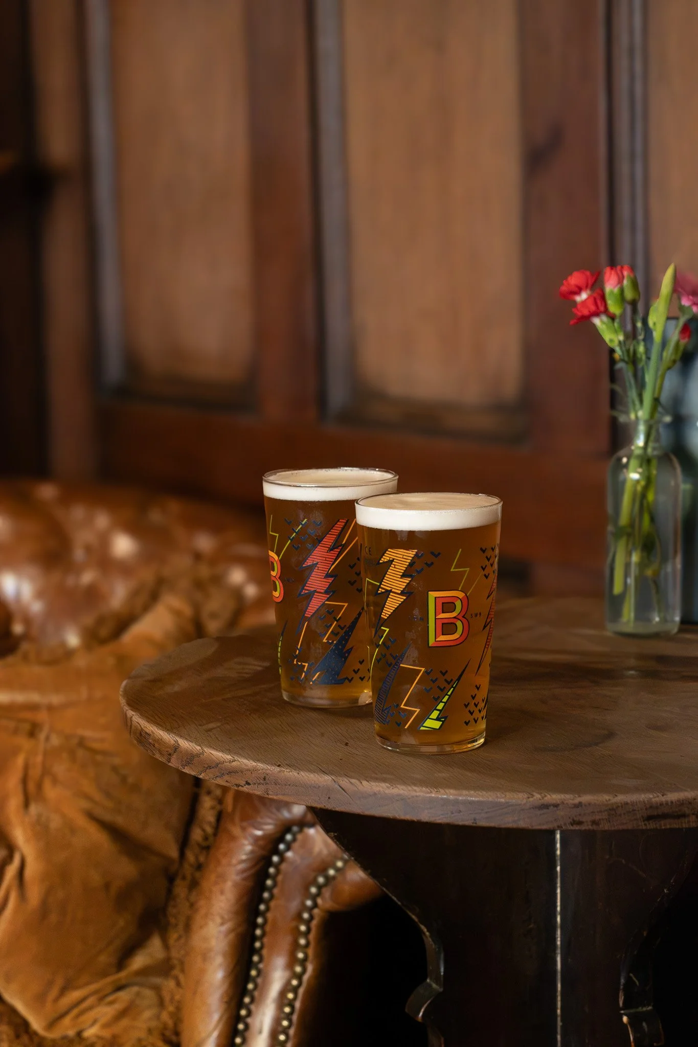 Two glasses of beer on a wooden table with a leather chair and a vase of red flowers in the background.  The Wilton Arms.