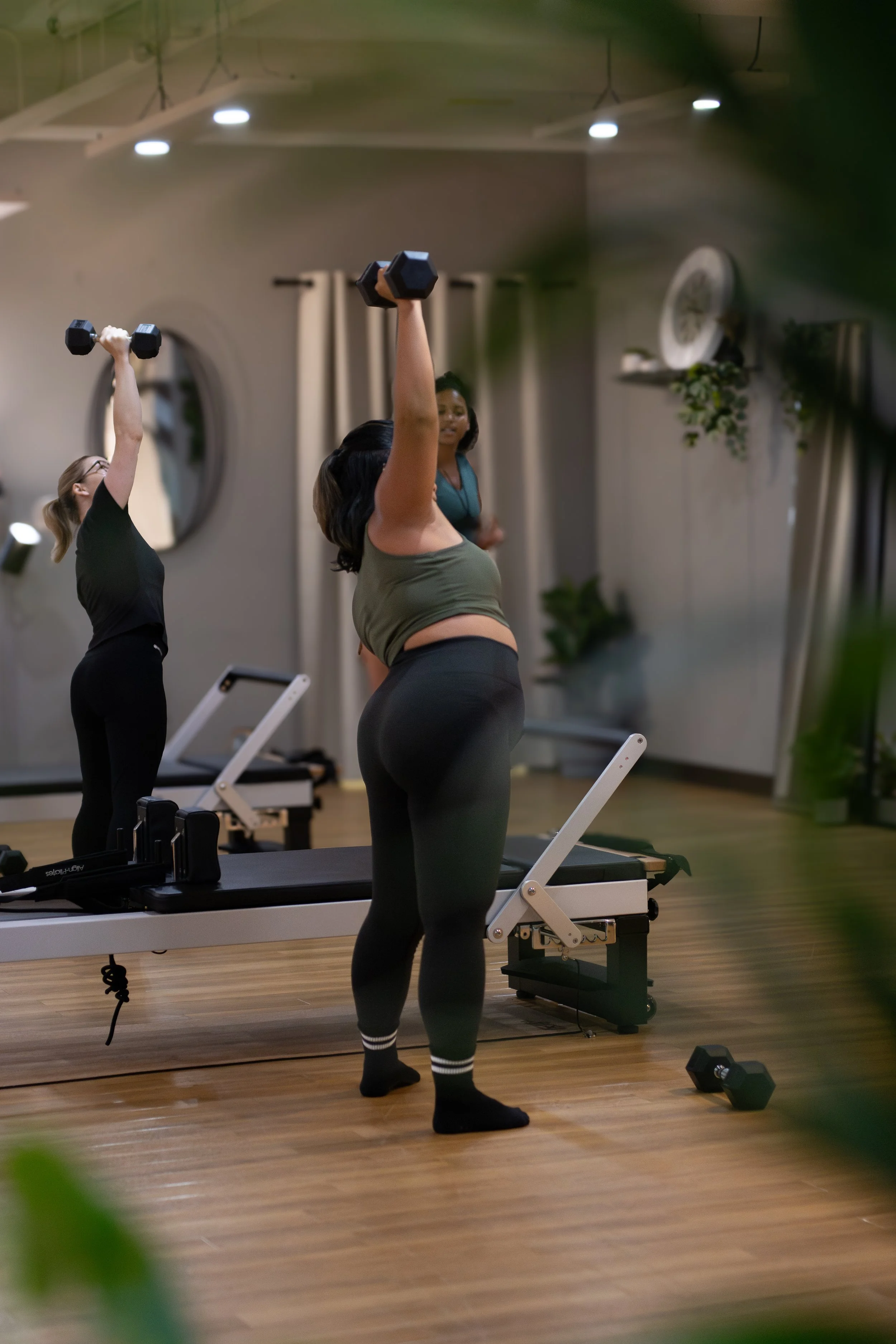 Two women lifting dumbbells during a workout in a gym, with exercise machines and wall decor visible in the background. The Studio Cheltenham, Reformer Pilates. 