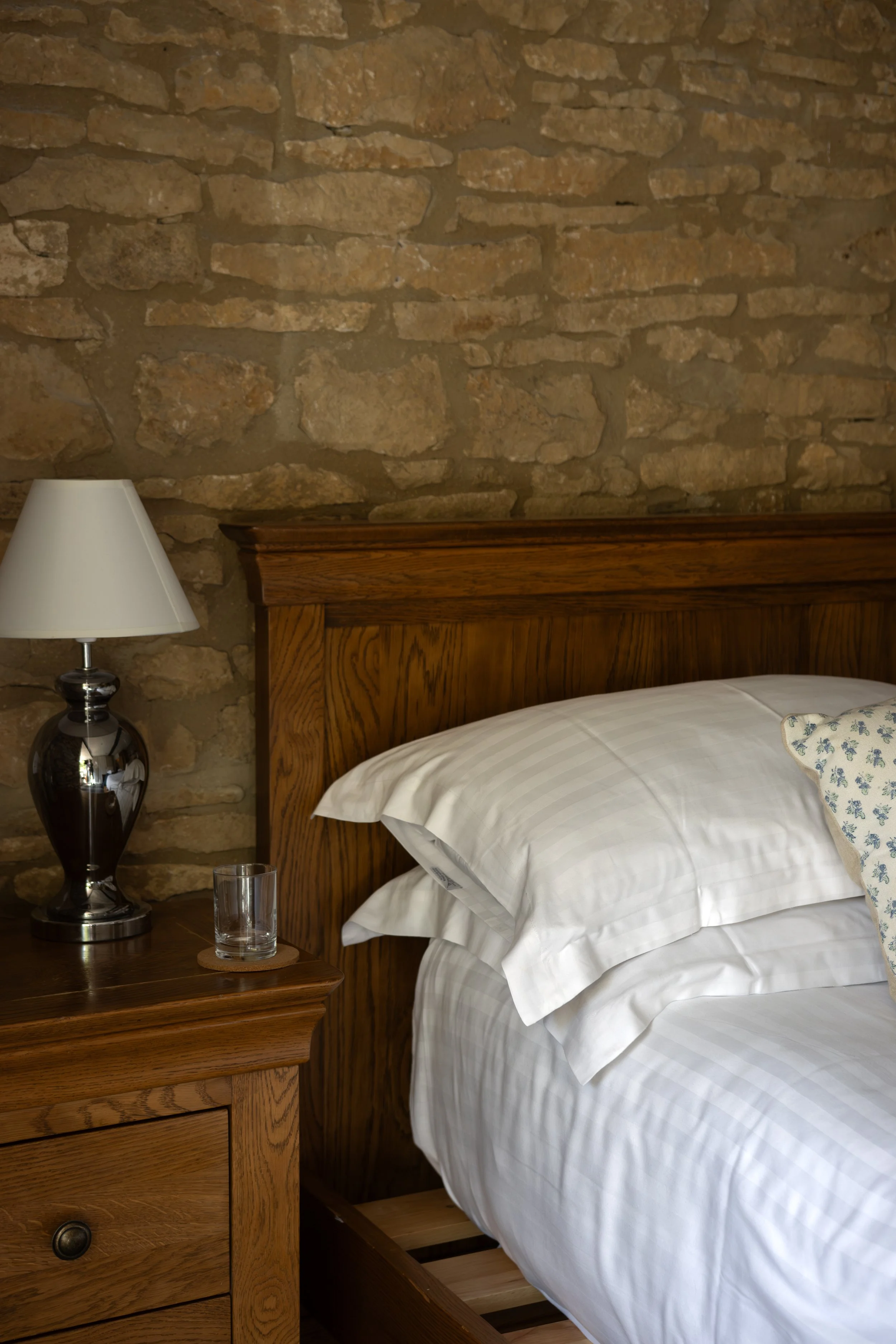 A close-up of part of a bedroom with a stone wall behind a wooden bed frame, a wooden nightstand, a lamp with a dark shiny base, a glass of water on a coaster, and a bed with white pillows and bedding with subtle stripes. The Lamb Inn. 