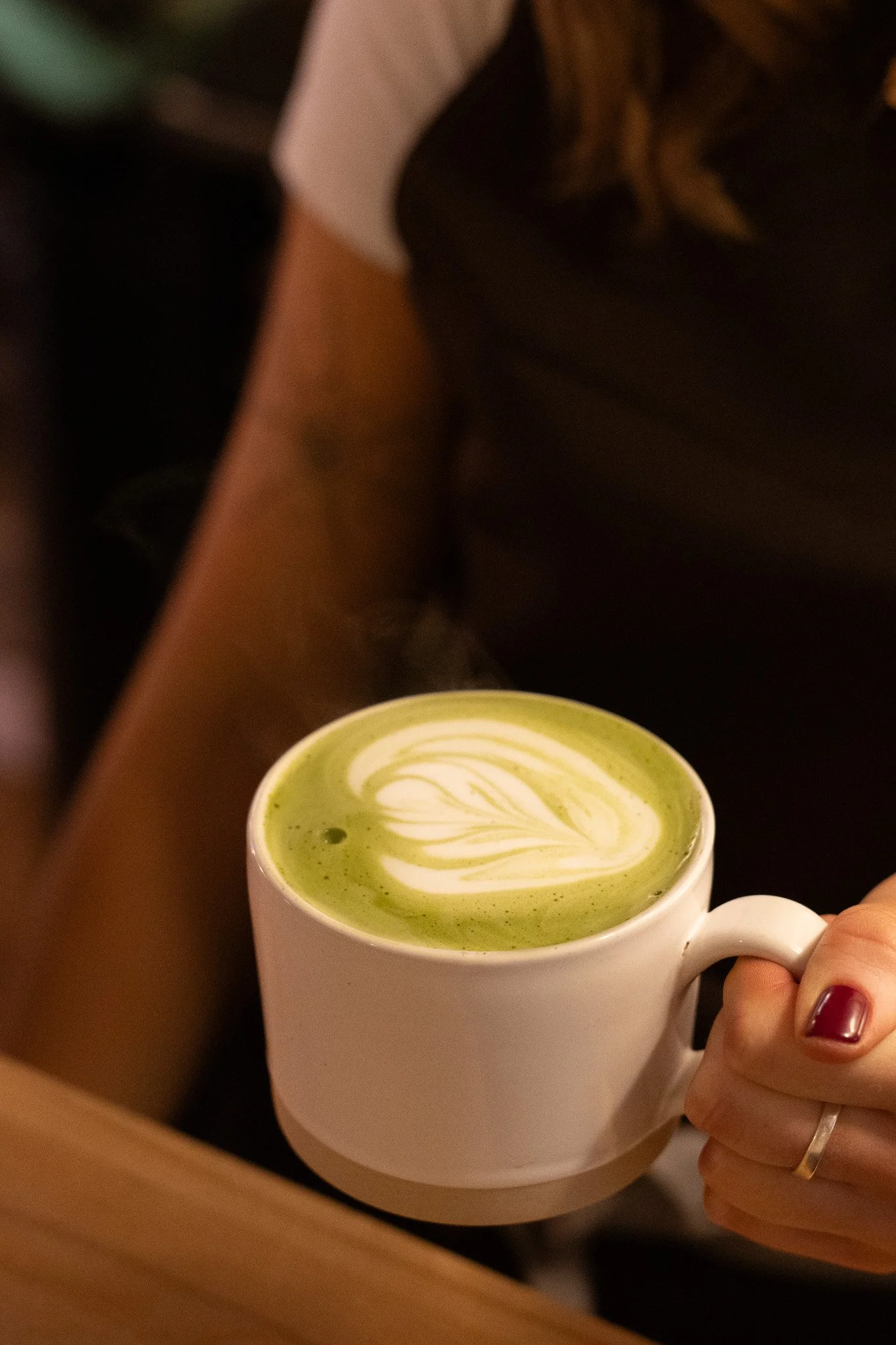 A person holding a white mug filled with green matcha latte with latte art. The New England Coffee House.
