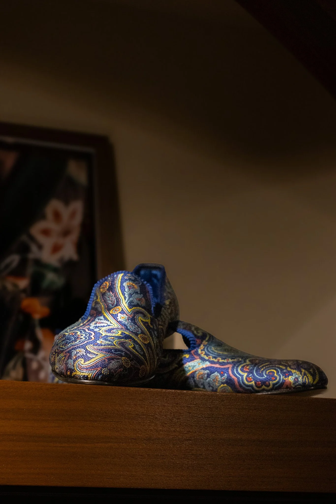 Close-up of a colorful paisley-patterned glass slipper on a wooden surface, with a blurred background. New & Lingwood.
