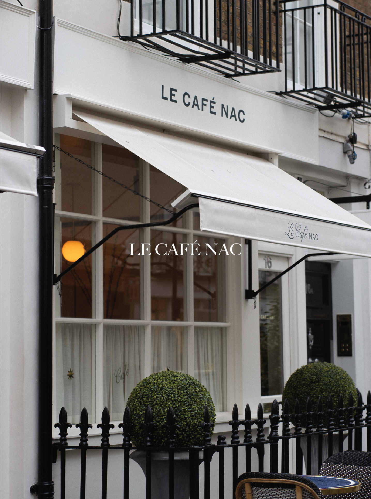 The exterior of Le Café NAC with a white storefront, black wrought iron railing fencing, potted topiary bushes, a window with white curtains, and a small round table with a yellow trim and wicker chairs outside. Le Cafe NAC.