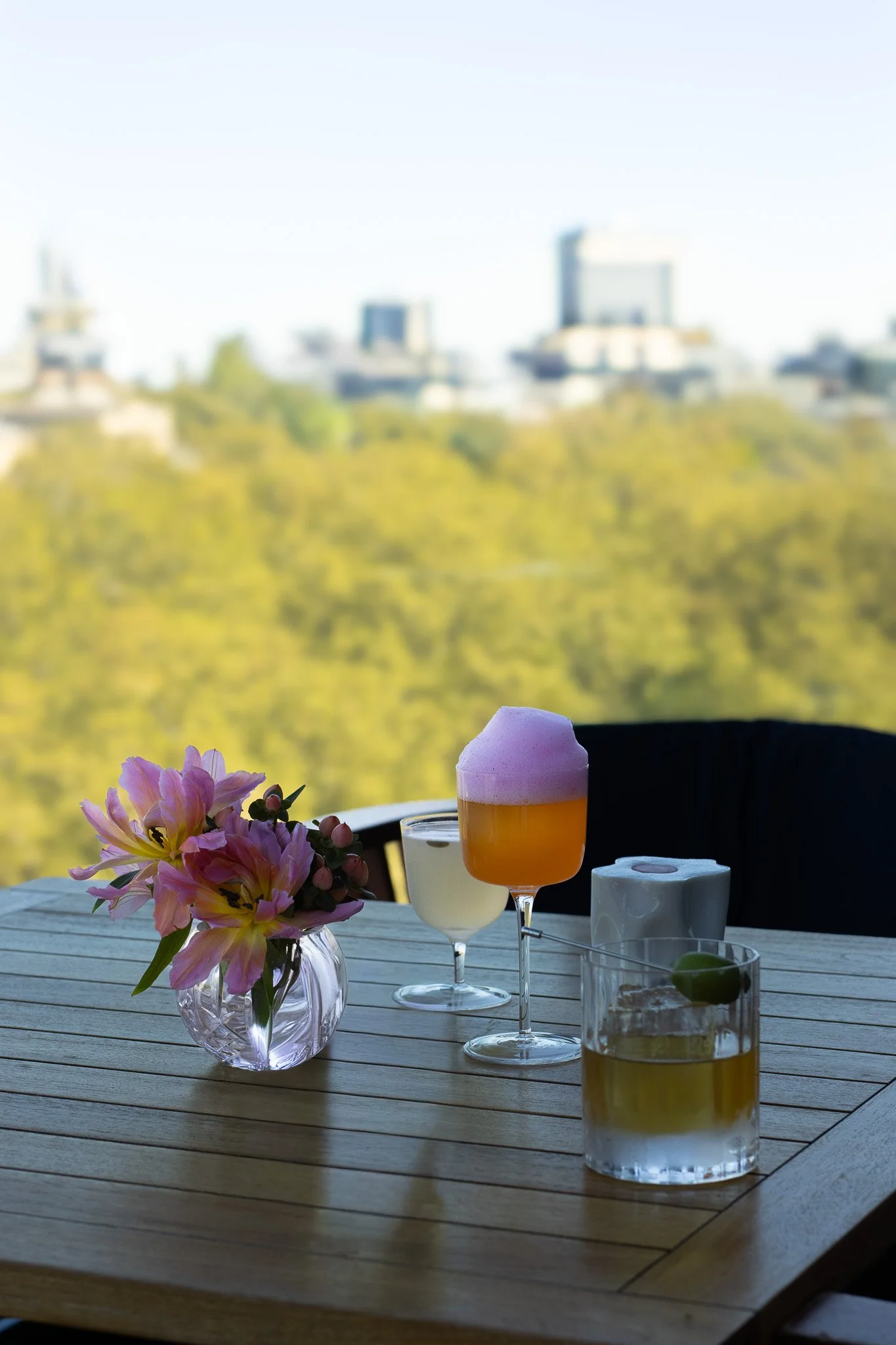 A wooden outdoor table decorated with a pink flower bouquet, three cocktails with foam and garnishes, and a white candle in a glass container, set against a scenic view of green trees and buildings in the distance. The Peninsula.