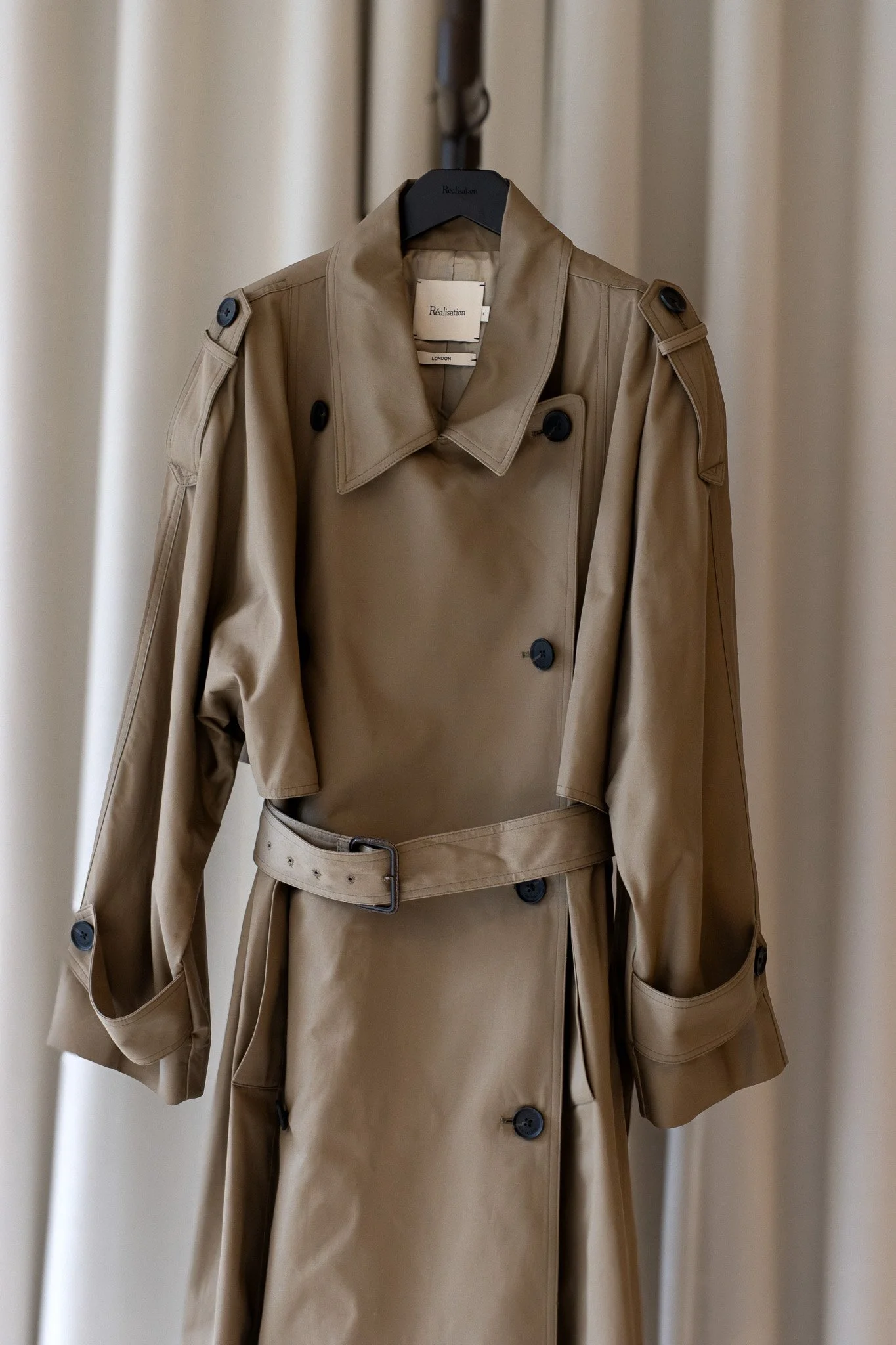 A beige trench coat hanging on a black hanger in front of a light-colored curtain. Realisation Par.