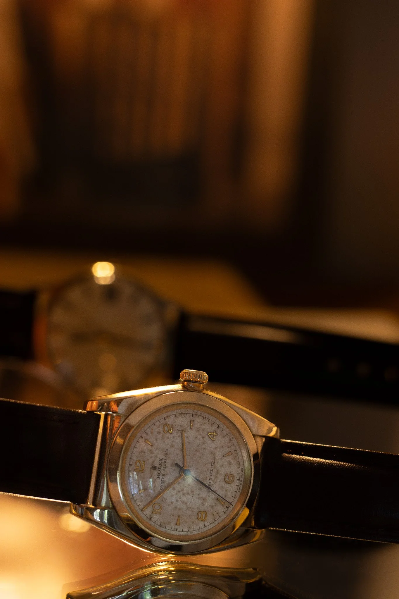A wristwatch with a white dial, gold case, black leather strap, and gold crown, placed on a shiny surface, with a blurred background of warm ambient lighting. Aurmont. Watches.