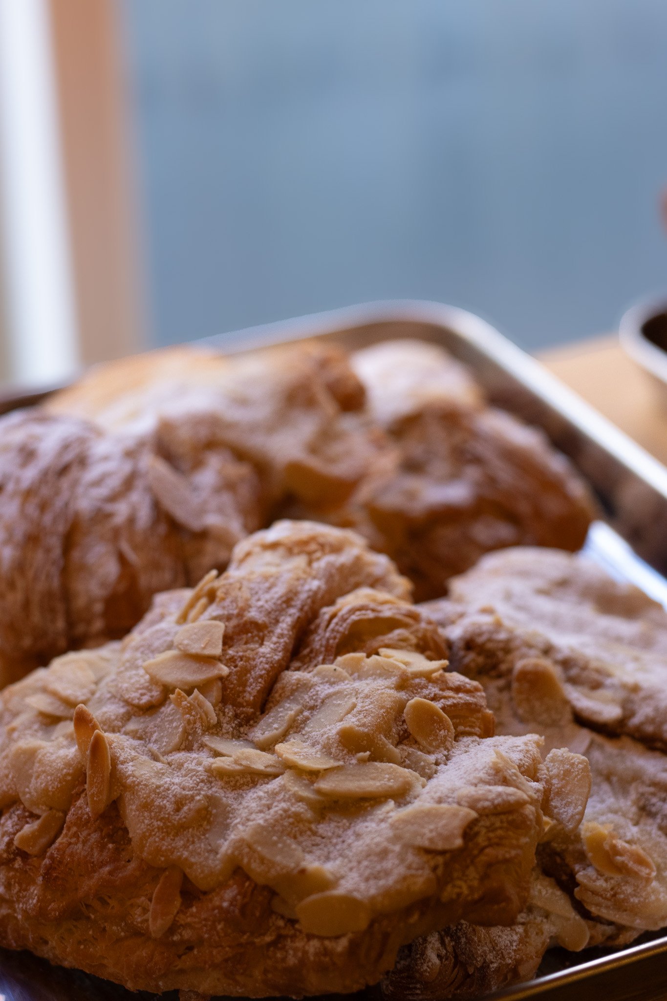 Close-up view of an almond pastry with sliced almonds on top, dusted with powdered sugar, on a baking sheet. The New England Coffee House.