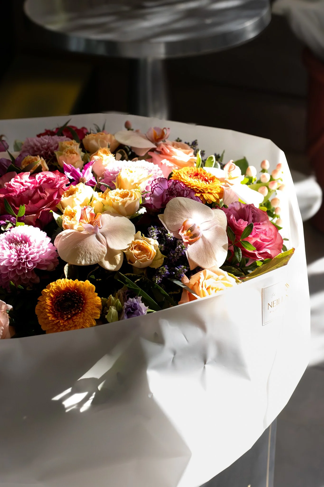 A large white floral bouquet with various colorful flowers, including roses, chrysanthemums, and orchids, wrapped in white paper. Neill Strain, Harrods, Motcomb Street.
