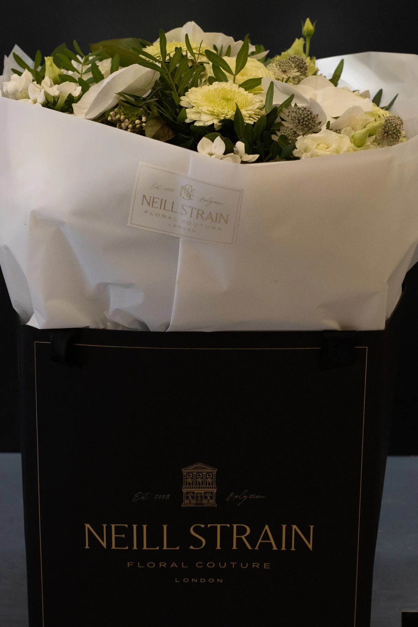 A bouquet of white flowers with greenery wrapped in white paper, labeled with a tag from Neil Strain Floral Couture London, placed on top of a black box with gold details. Neill Strain, Harrods, Motcomb Street.
