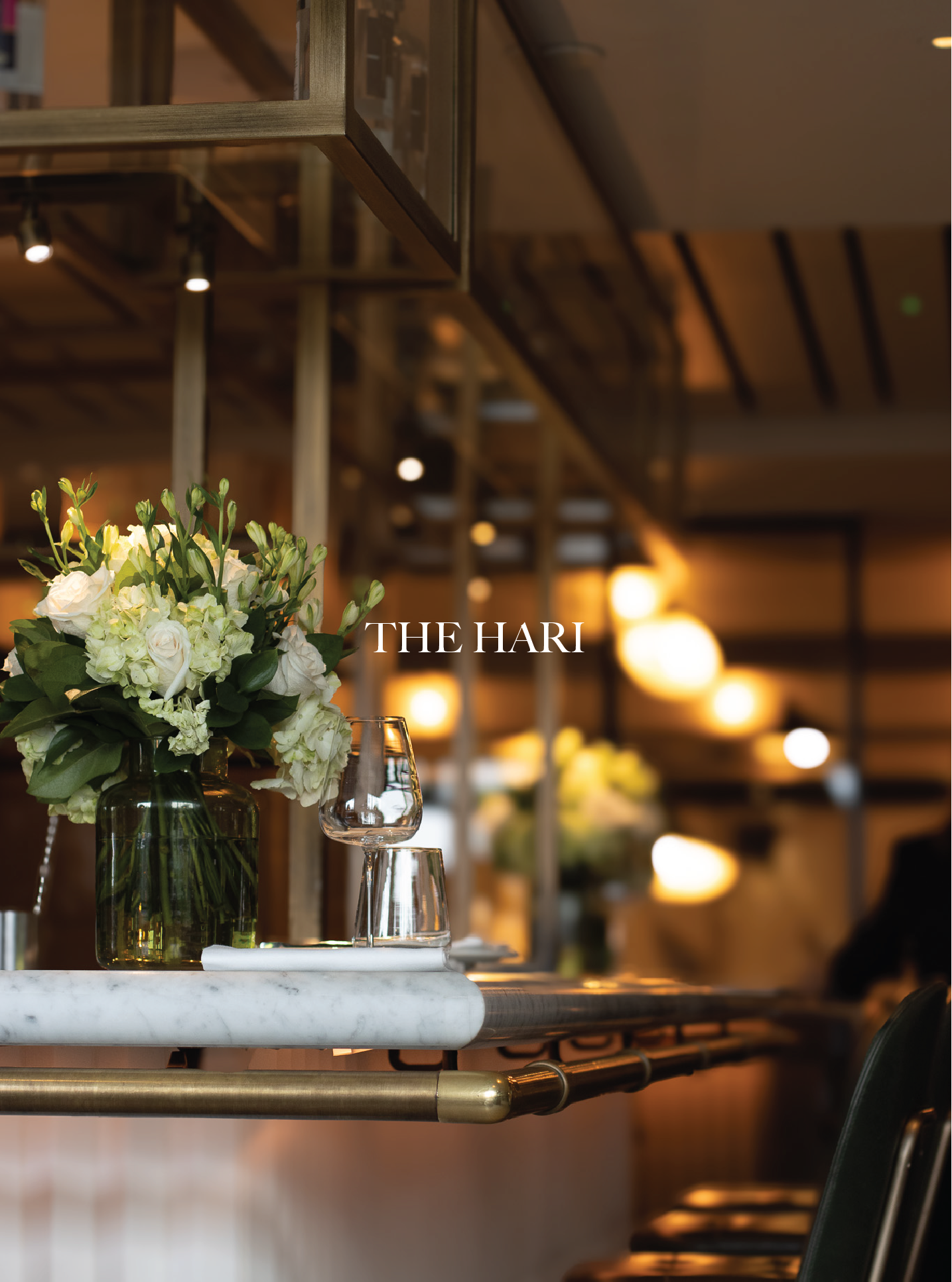 A floral arrangement with white and green flowers, placed on a marble countertop with wine glasses nearby. The background shows warm, dim lighting in a restaurant or event space with modern decor. The Hari, Knightsbridge, Il Pampero.