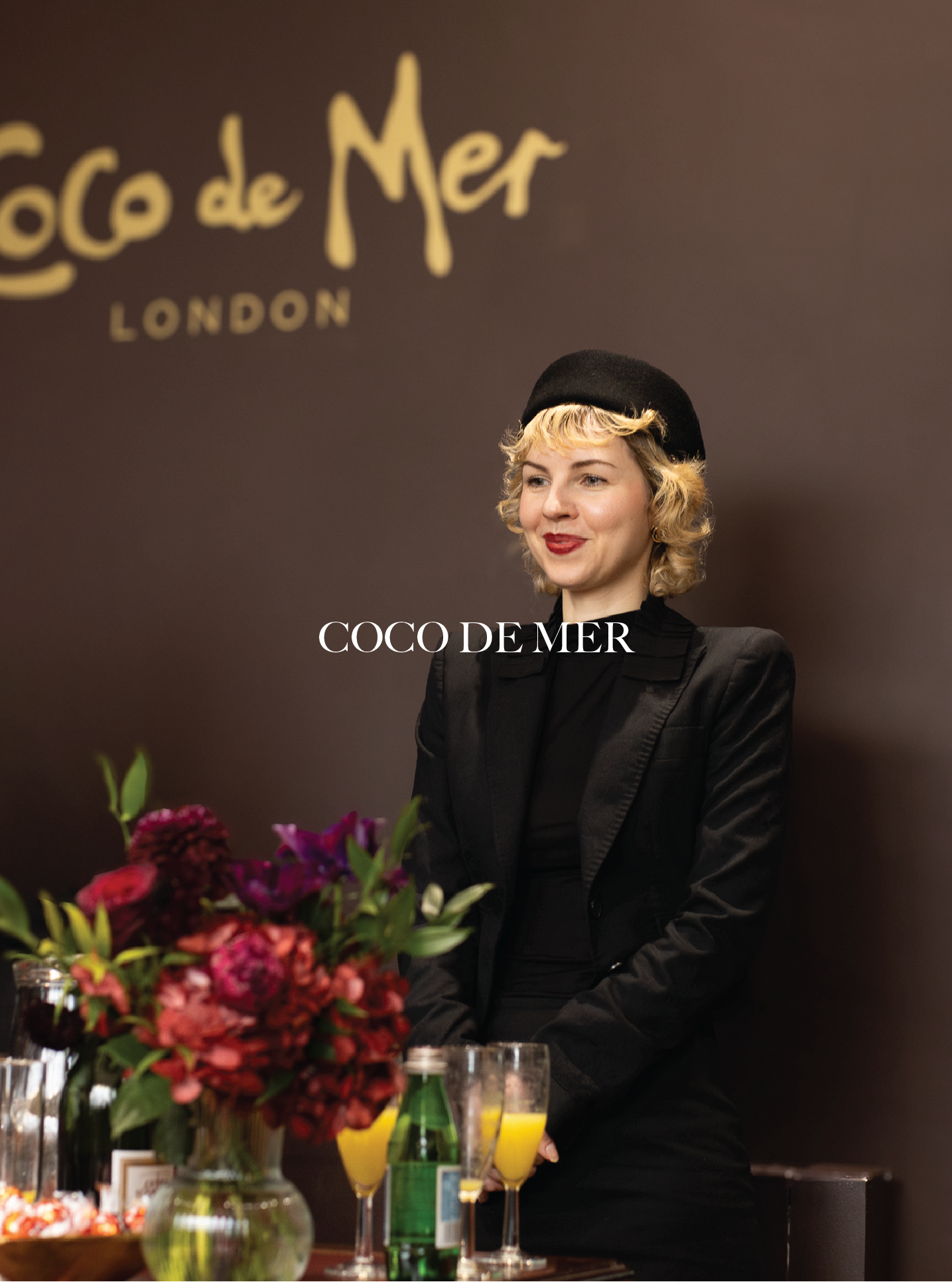 A woman with blonde curly hair wearing a black beret and black blazer standing at a table with flowers and drinks, with a dark background featuring the 'Coco de Mer London' logo.
