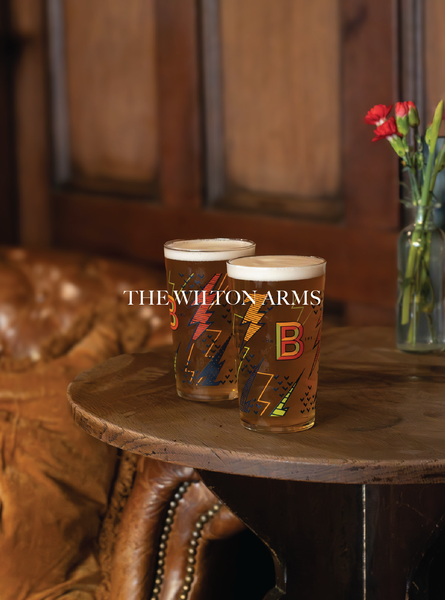 Two pint glasses of beer on a round wooden table, with a bouquet of red flowers in a glass vase in the background. The setting appears to be a cosy bar or pub with wood-paneled walls. The Wilton Arms Pub.