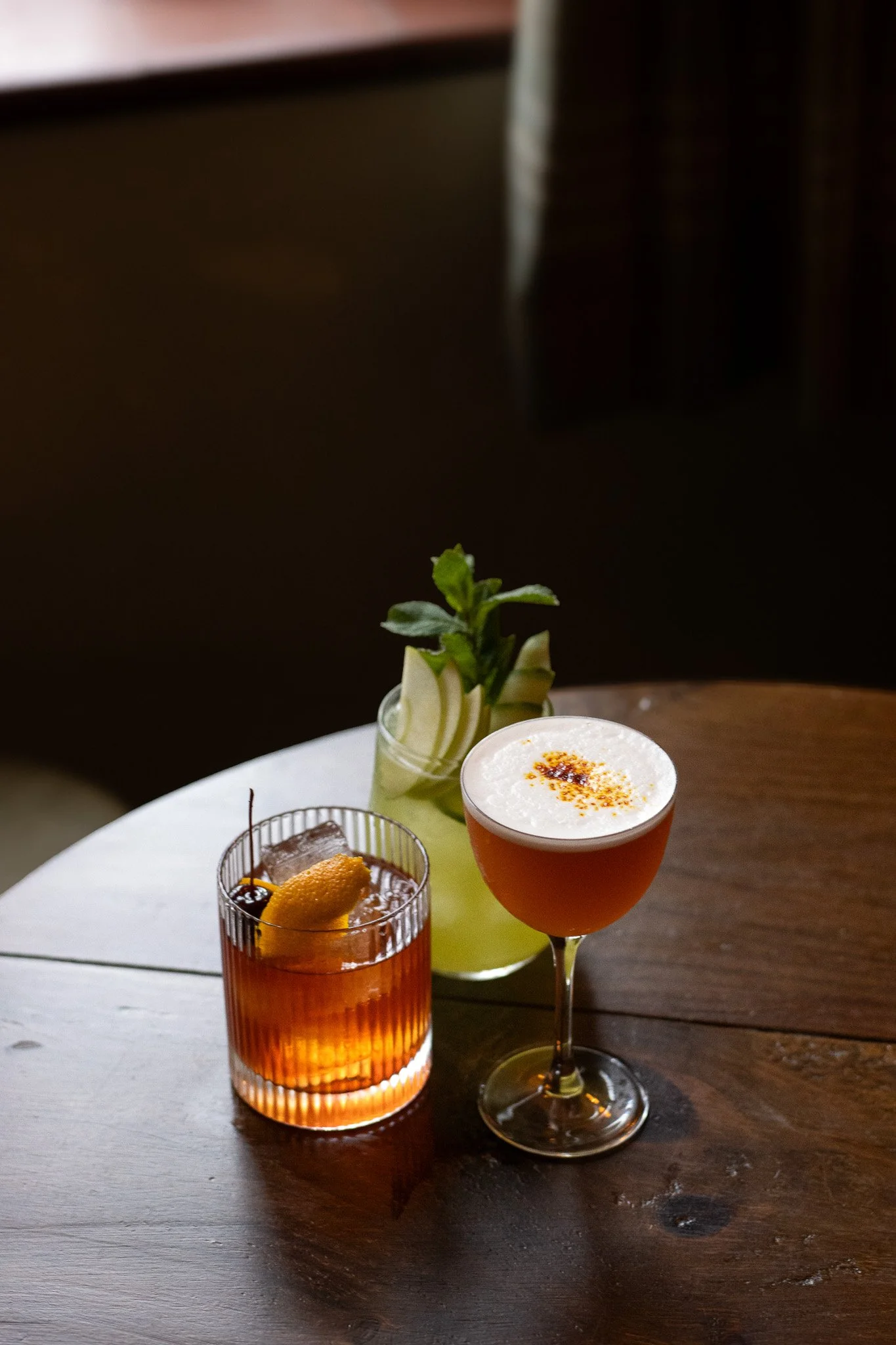 Three cocktails on a round table: a blue drink with orange peel, an amber drink with cherry, and a pink cocktail with frothy top and spices, with a plant in the background. The Lamb Inn. 
