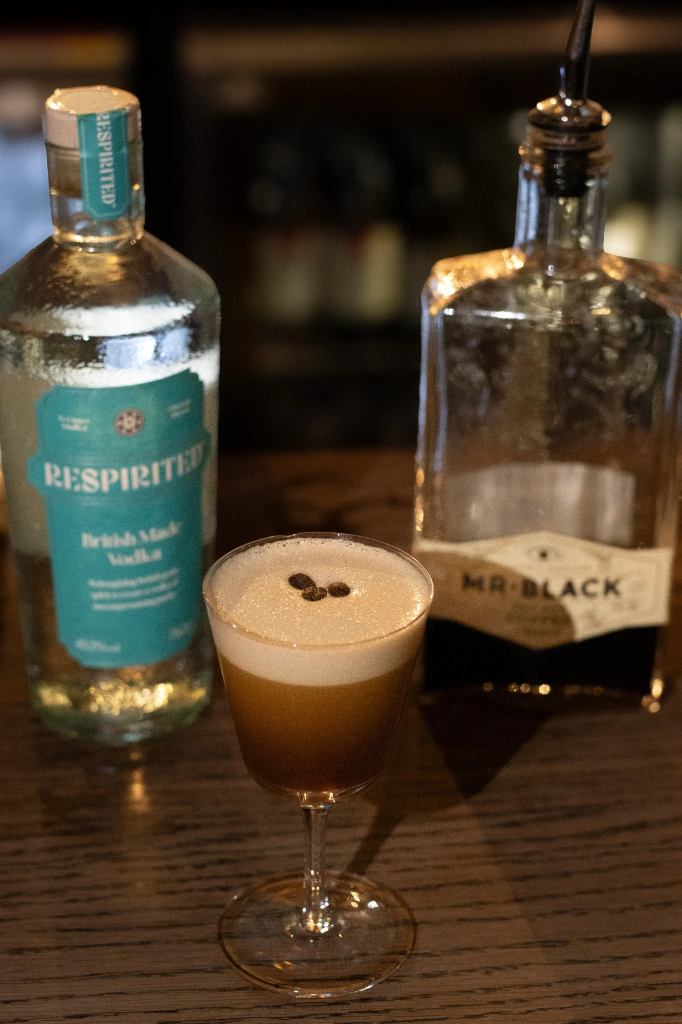 A cocktail topped with coffee beans in front of a bottle of ReSpirit and a bottle of Mr. Black Nero Coffee Liqueur on a wooden table. Alfred Tennyson.