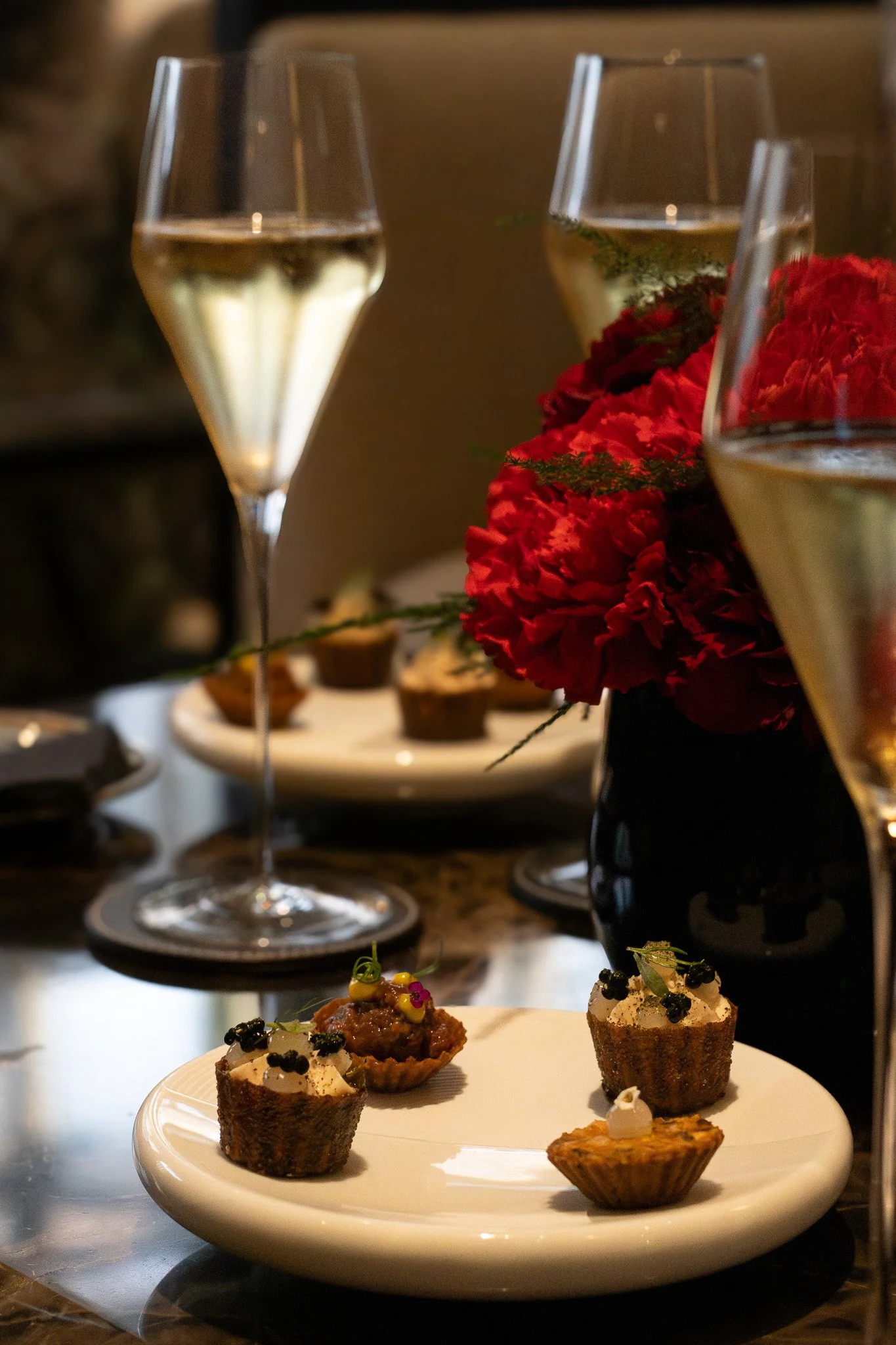 Two glasses of white wine, a red flower arrangement, and a plate of assorted appetisers on the table. Petrus.