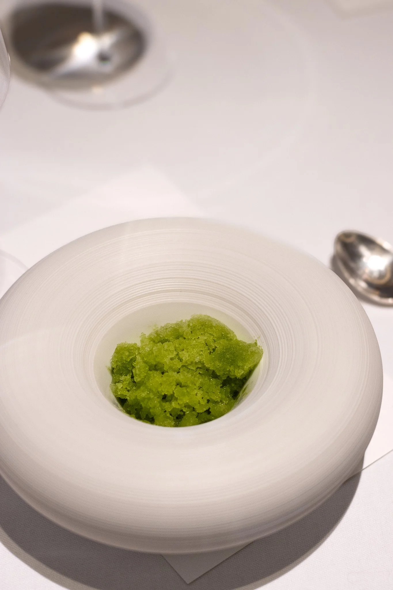 A white bowl containing green sorbet or ice cream on a white surface, with metal salt and pepper shakers visible in the background. Petrus.