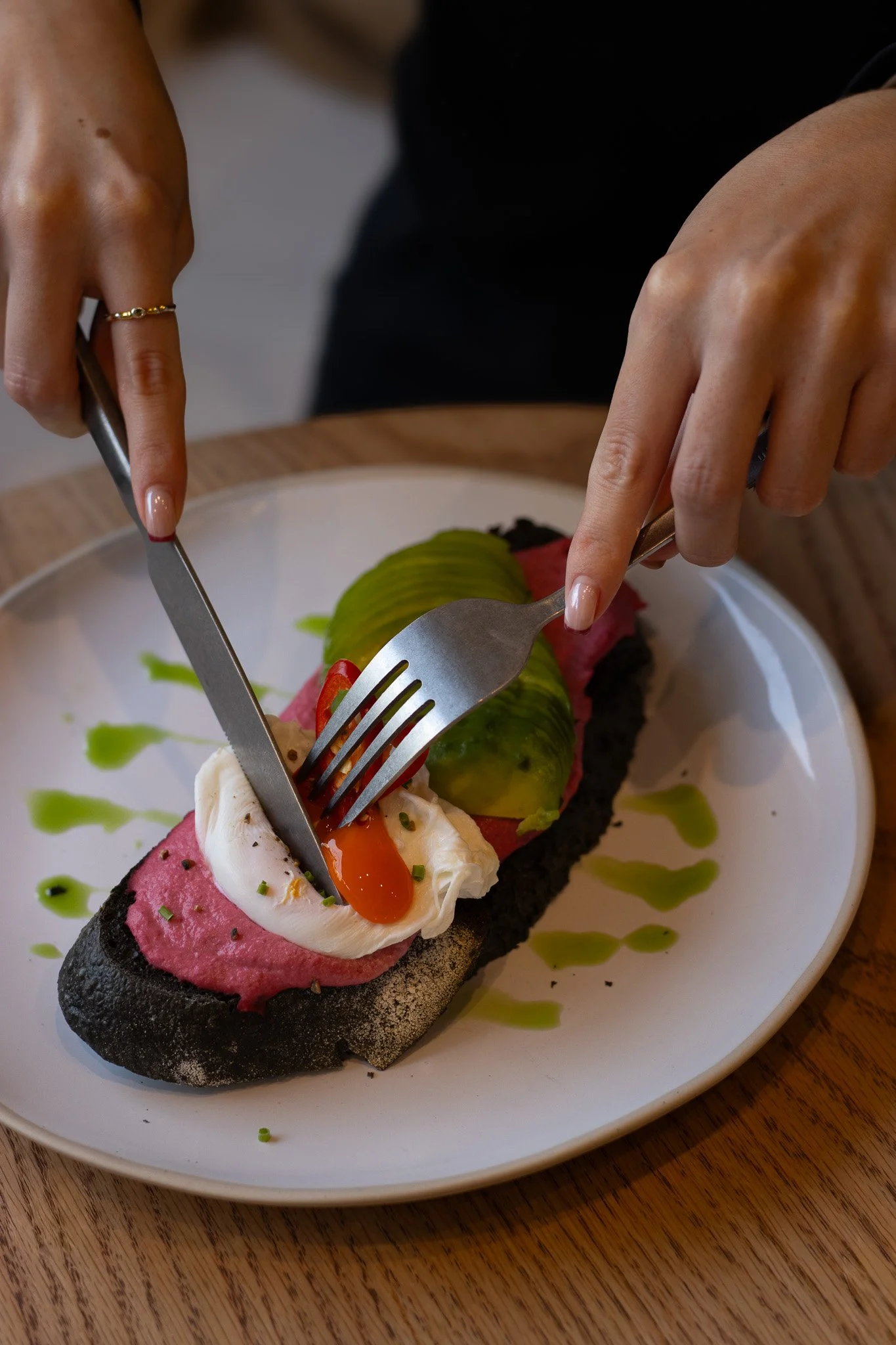 A slice of charcoal bread topped with pink pâté, a soft-poached egg, a slice of cucumber, and red chili slices, garnished with chopped herbs and presented on a white plate with green sauce drizzled around. The Cotswold Farm. 