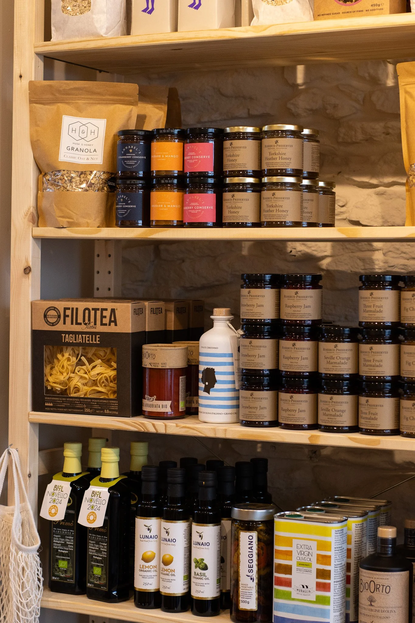 Wooden shelf containing jars of jam, honey, and preserves, alongside boxes of pasta and bottles of olive oil in a pantry or kitchen storage area. The Cotswold Farm. 
