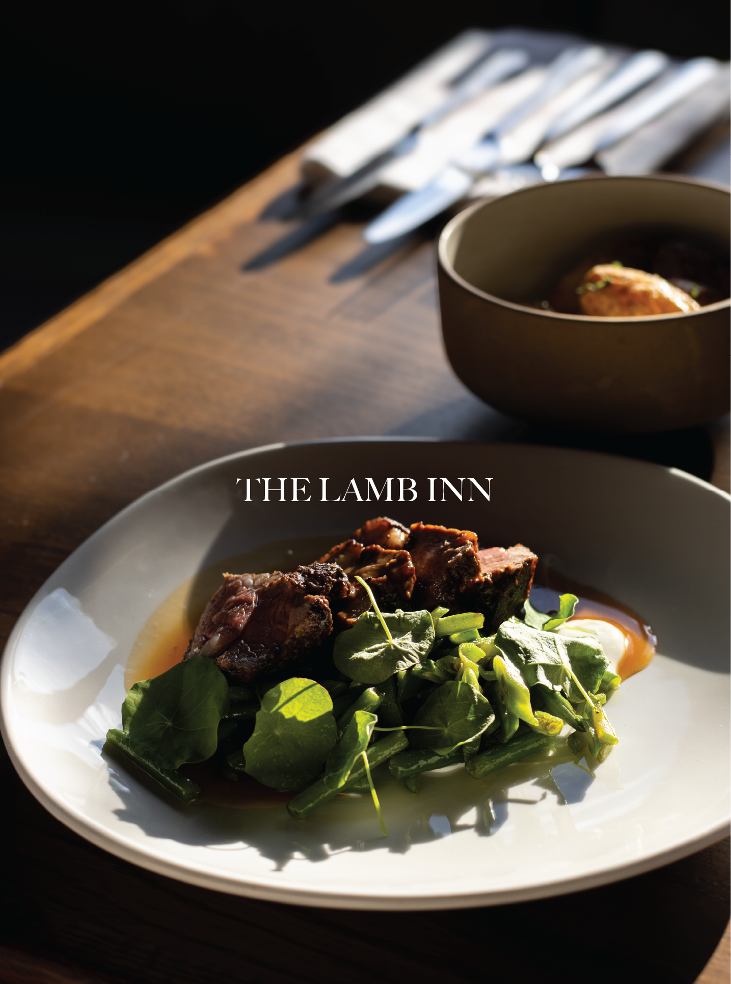 A white plate with a serving of roasted beef with sauce, accompanied by greens. In the background, a black bowl with potatoes and a wooden table with silverware. The Lamb Inn, Cotswolds.