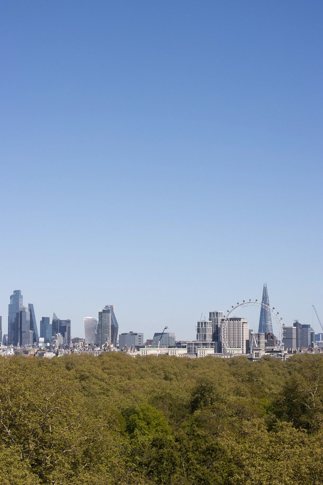 London skyline with trees in the foreground, including the Shard and the London Eye, under a clear blue sky. The Peninsula.