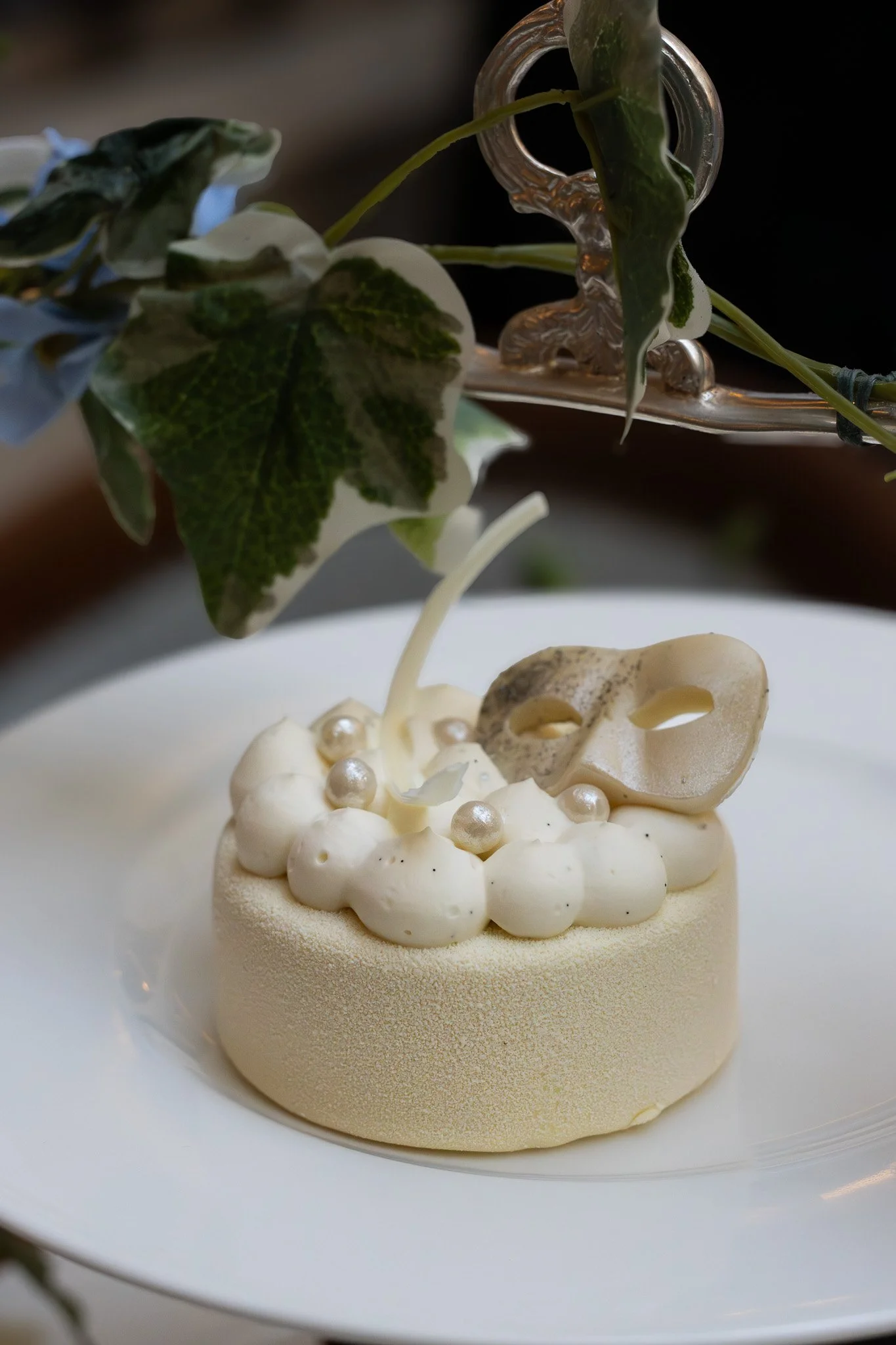 A white, round dessert topped with white cream or mousse, decorated with small edible pearls. It is served on a white plate, with green leaves and a silver decorative item in the background. The Lanesborough.