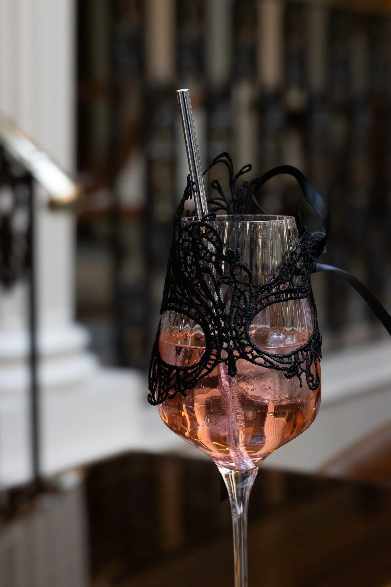 A glass of pink drink with black lace decoration around the rim, with a metal straw inside, set against a blurred background.  The Lanesborough. Bridgerton Afternoon Tea.