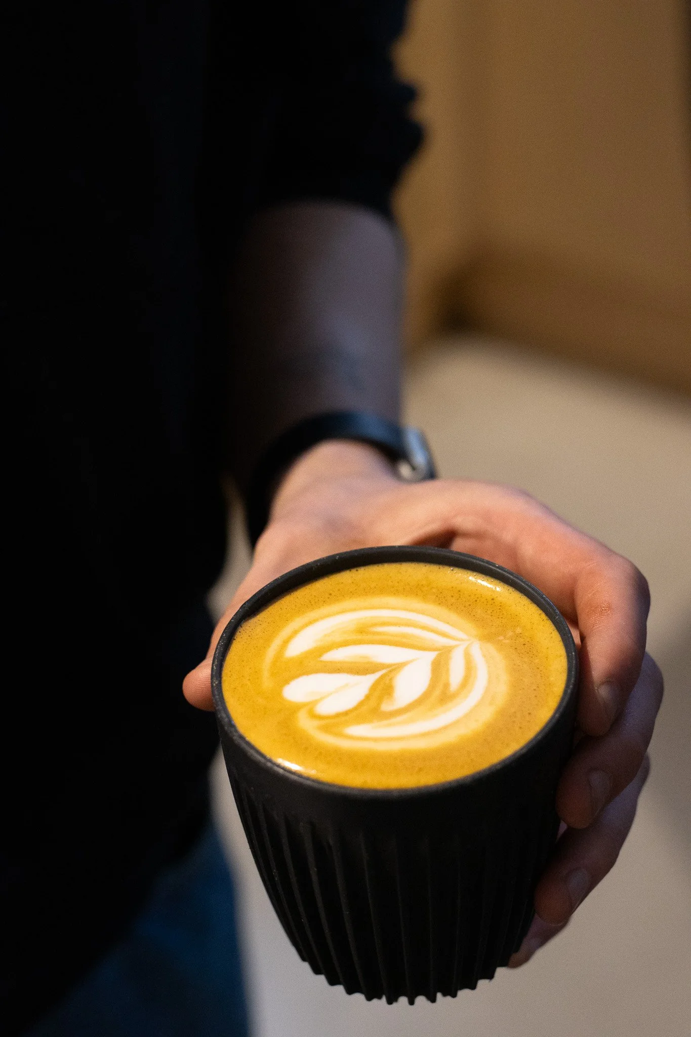 A person holding a black cup of coffee or latte with latte art in the shape of a leaf. The Scandinavian Coffee Pod.