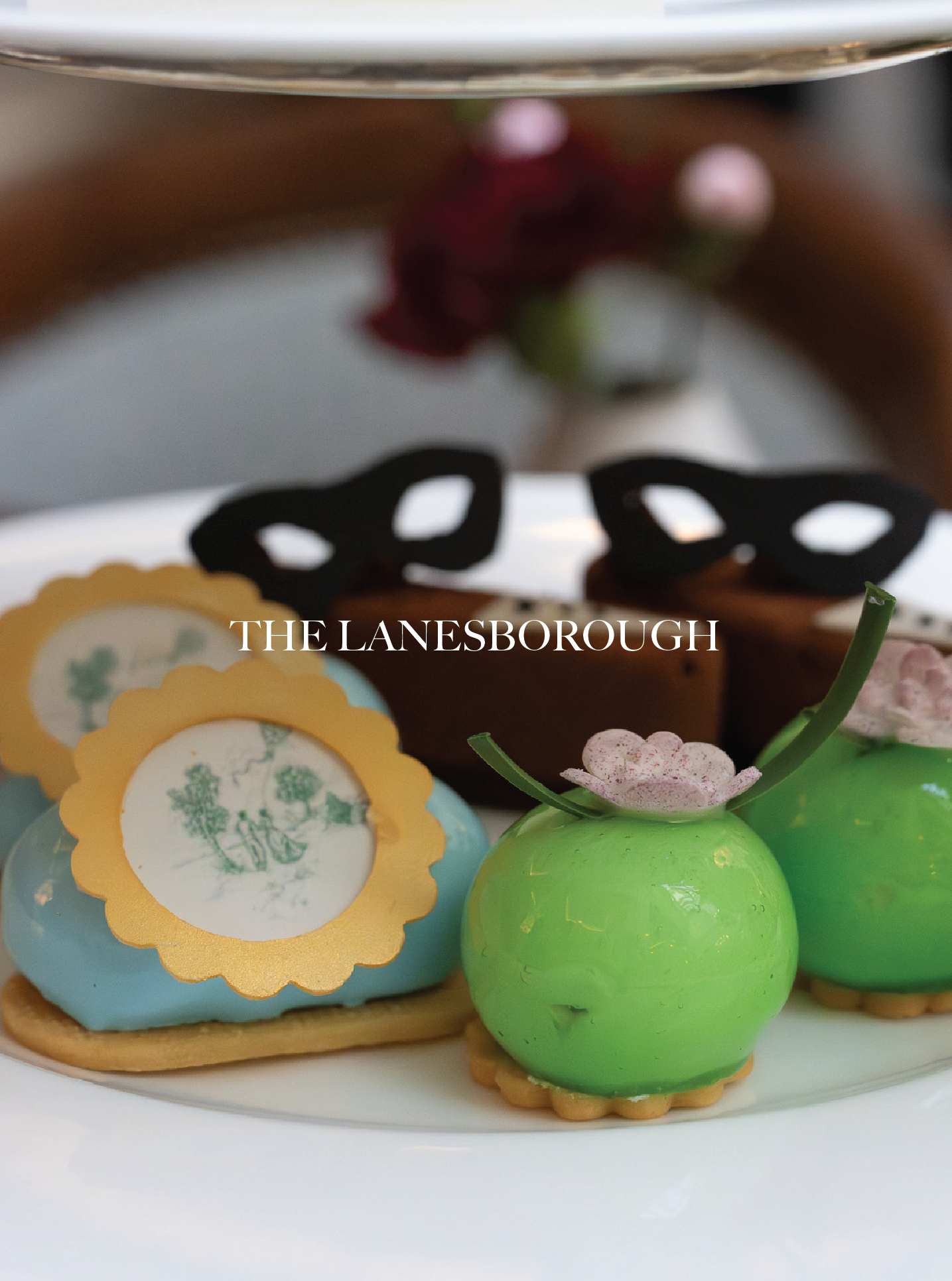 Colorful decorated Easter eggs with one in a bright green color, topped with a small pink and white flower, and another with a vintage picture design and a scalloped yellow border, displayed on a white plate. The Lanesborough.