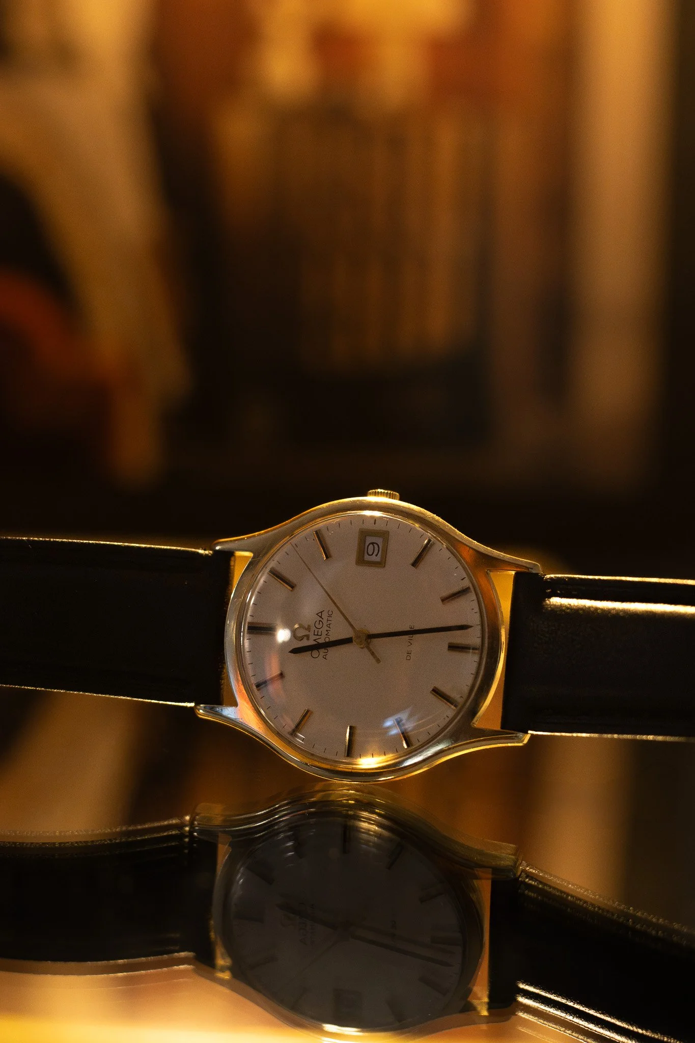 Close-up of a gold Omega automatic watch with a white face, black leather band, and visible date window, reflected on a shiny surface with blurred background. Aurmont. Watches.