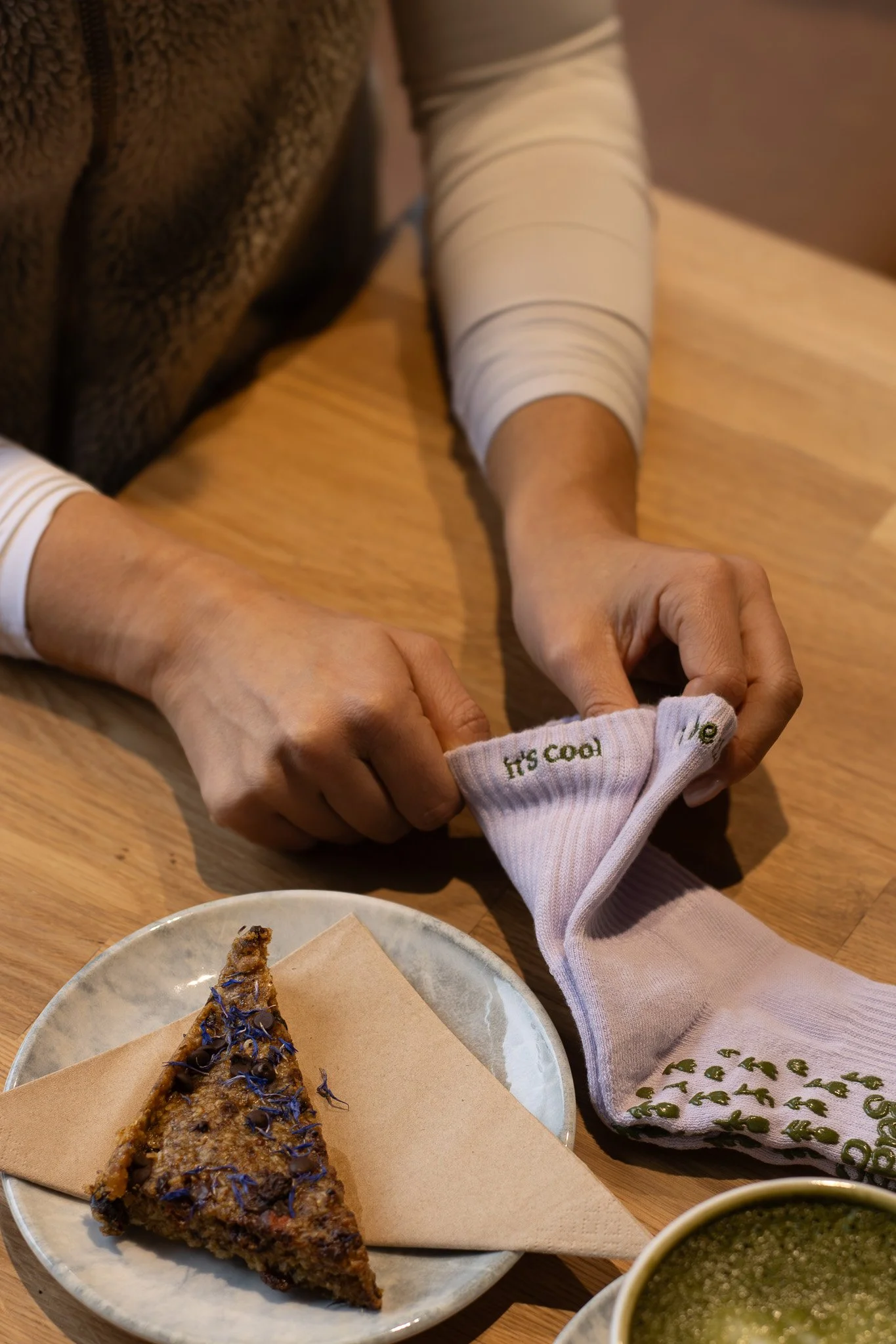 A person helping a young child put on their sock in a cozy setting with a wooden table, a plate with a slice of pie, and a cup of green beverage. GetAGrip Apparel. 