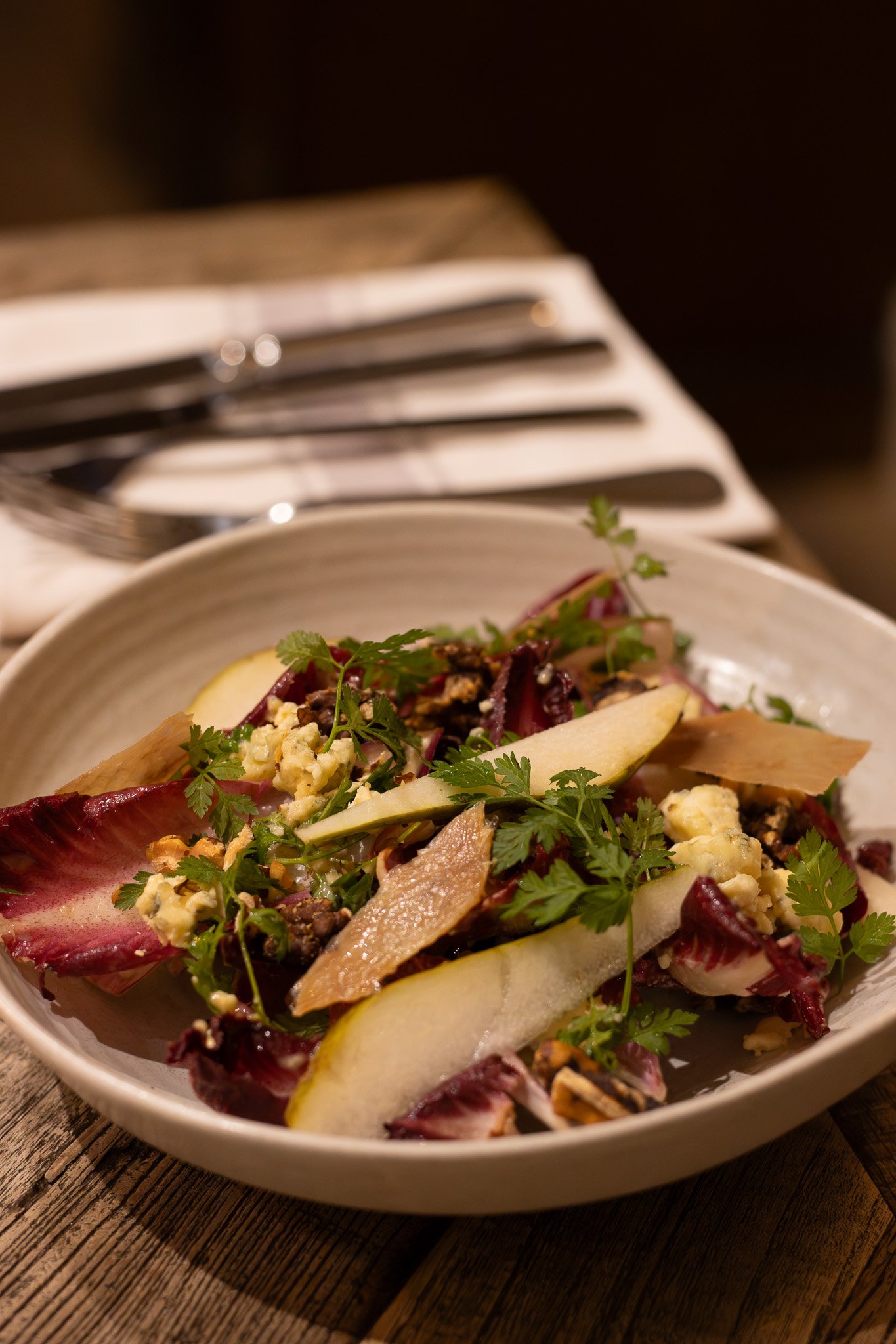 Close-up of a salad in a white bowl with sliced pears, beets, chopped nuts, cheese, and garnished with parsley on a wooden table. The Lamb Inn. 