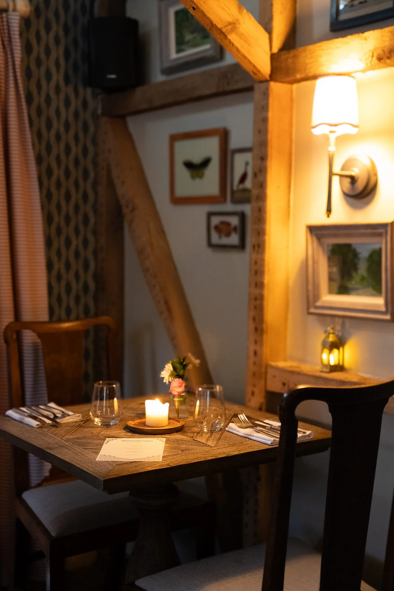 A cozy dining room with a wooden table set for a meal, featuring wine glasses, plates, and utensils. The table is decorated with candles and a small flower arrangement. The room has warm lighting from a wall sconce and candlelight, with framed pictur