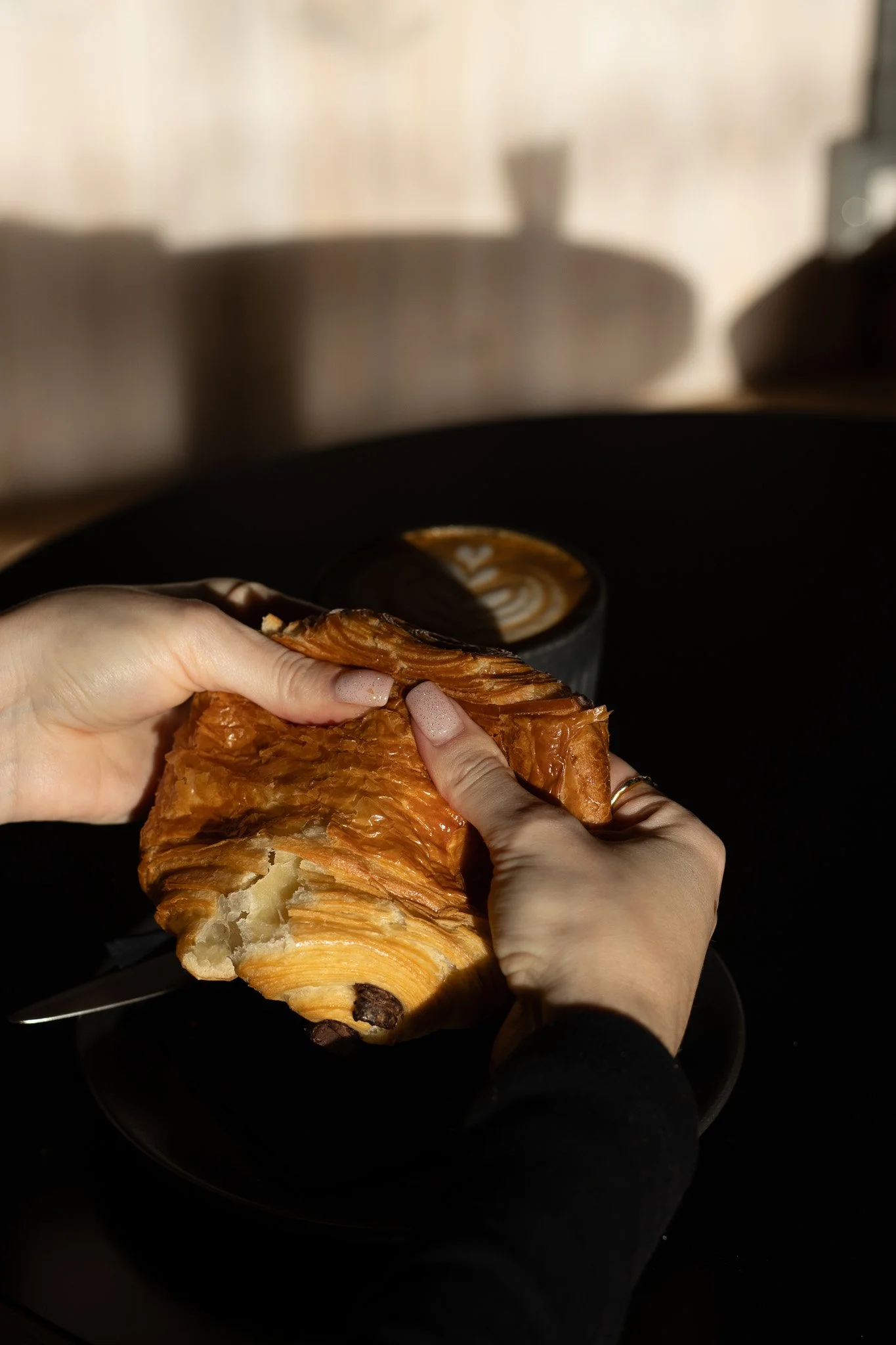 Person holding a piece of a chocolate croissant in front of a coffee cup on a black plate, with a blurred background. The Scandinavian Coffee Pod.