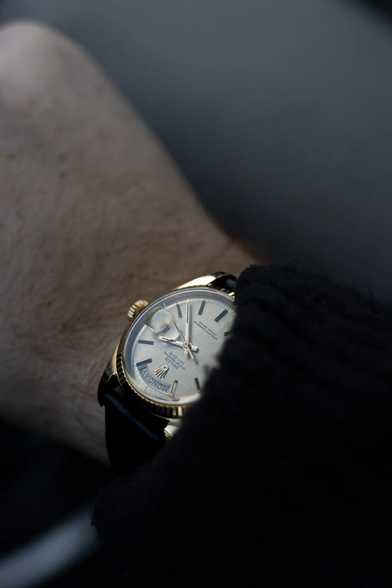 A person's wrist wearing a Rolex watch with a light-colored face, black leather strap, and gold bezel, viewed partially with a blurred background. Aurmont. Rolex. Watches.