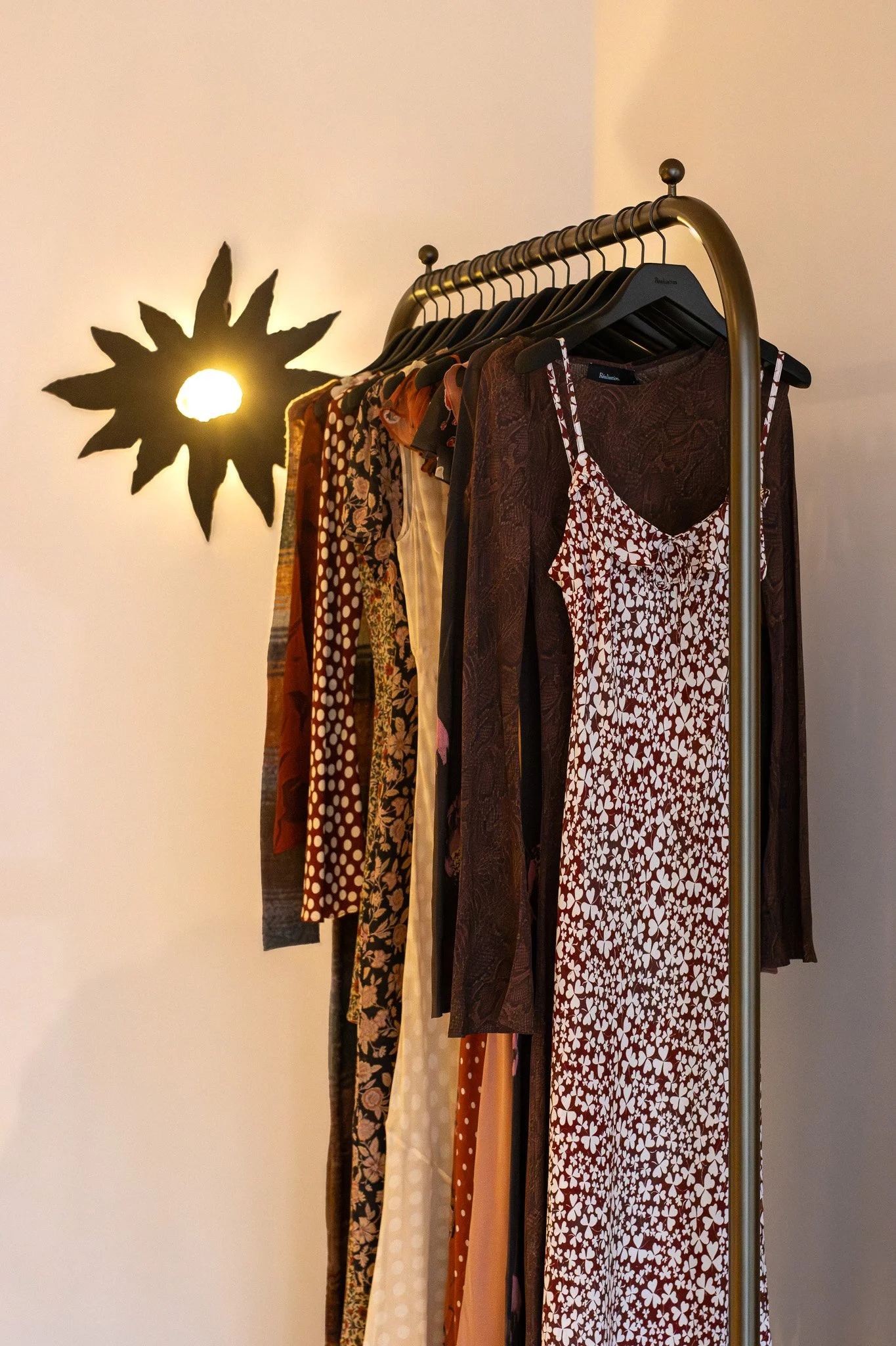 Clothing rack with various dresses and garments hanging, illuminated by a wall-mounted light fixture shaped like a sunflower. Realisation Par.