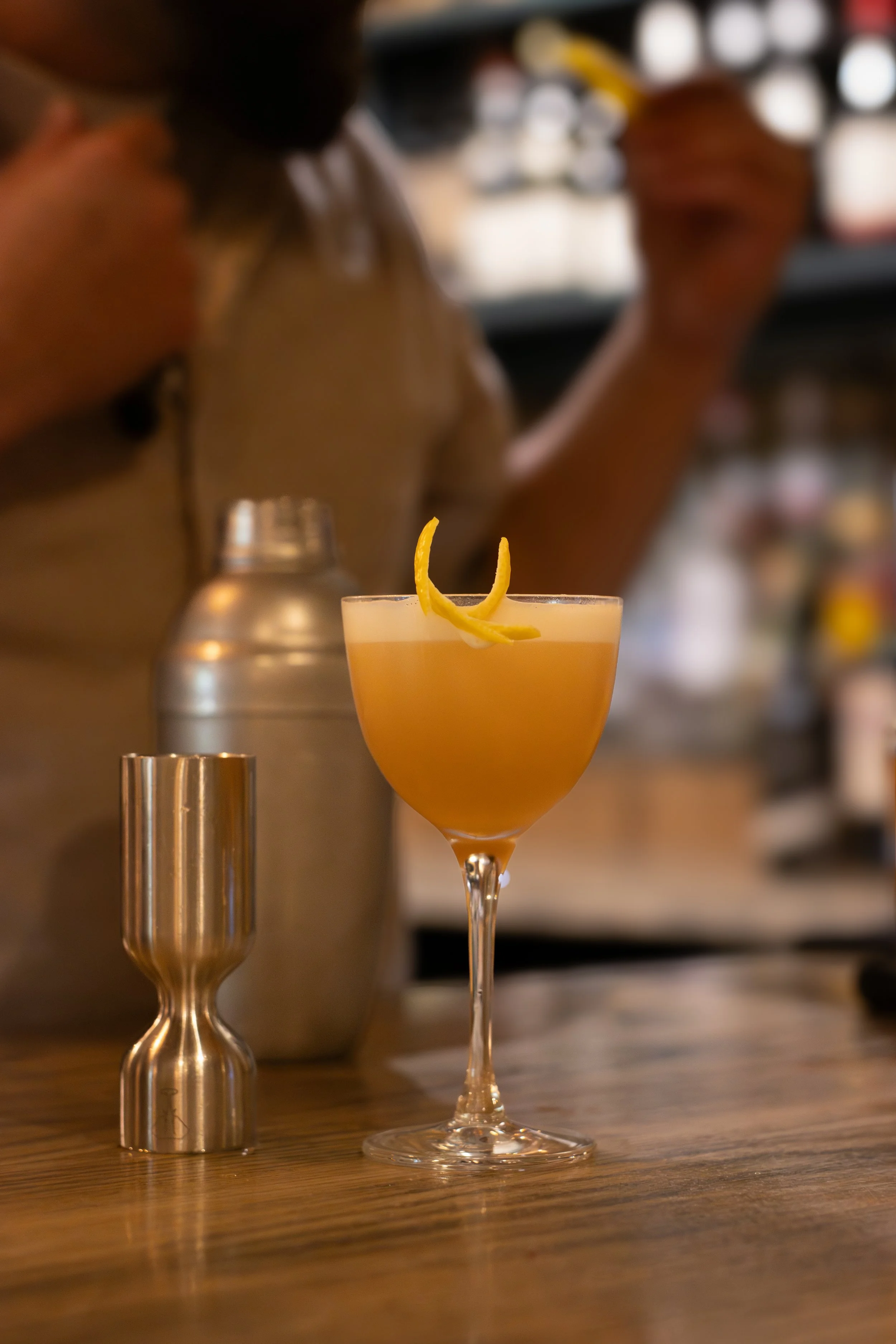 Close-up of a cocktail glass with a lemon twist garnish on a wooden bar counter, with blurred bartender in the background. The Lamb Inn. 
